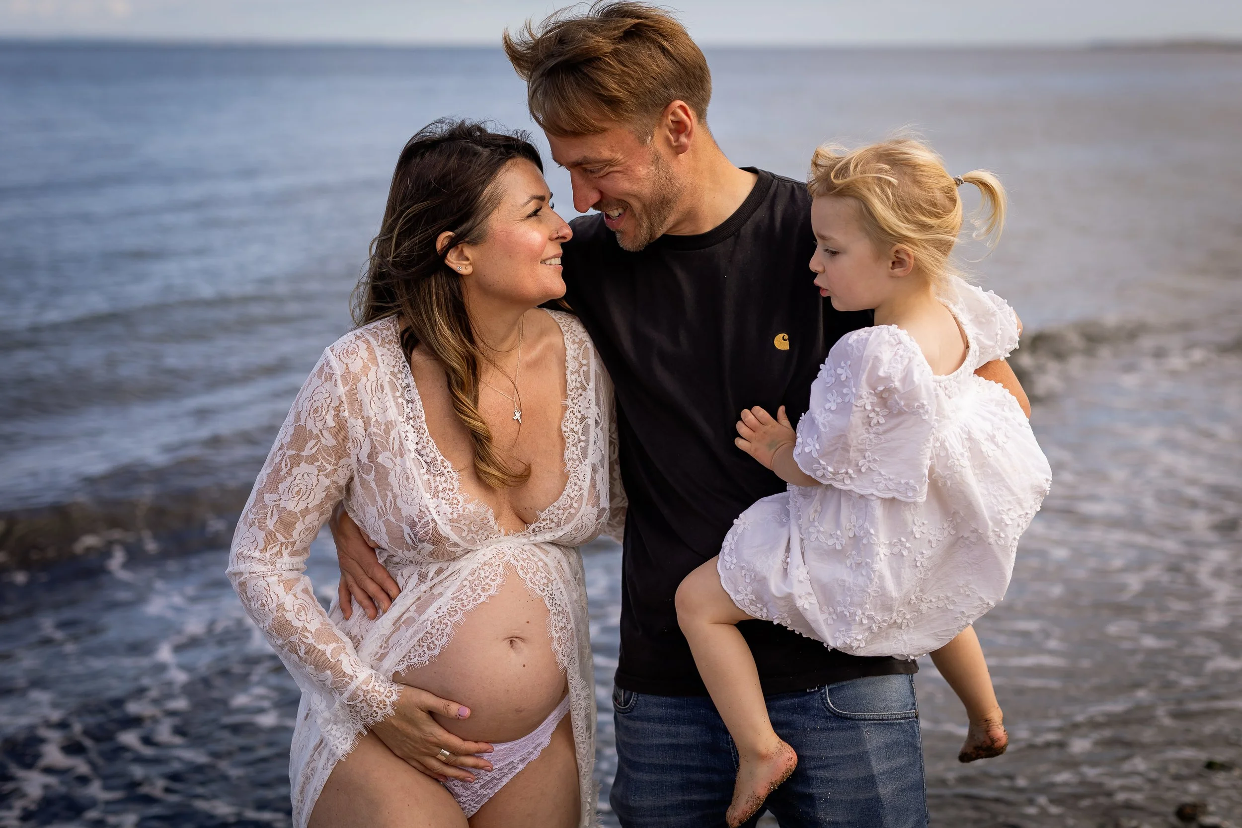 Pregnant mum in a white lace dress standing beside her partner as he holds their daughter by the sea.