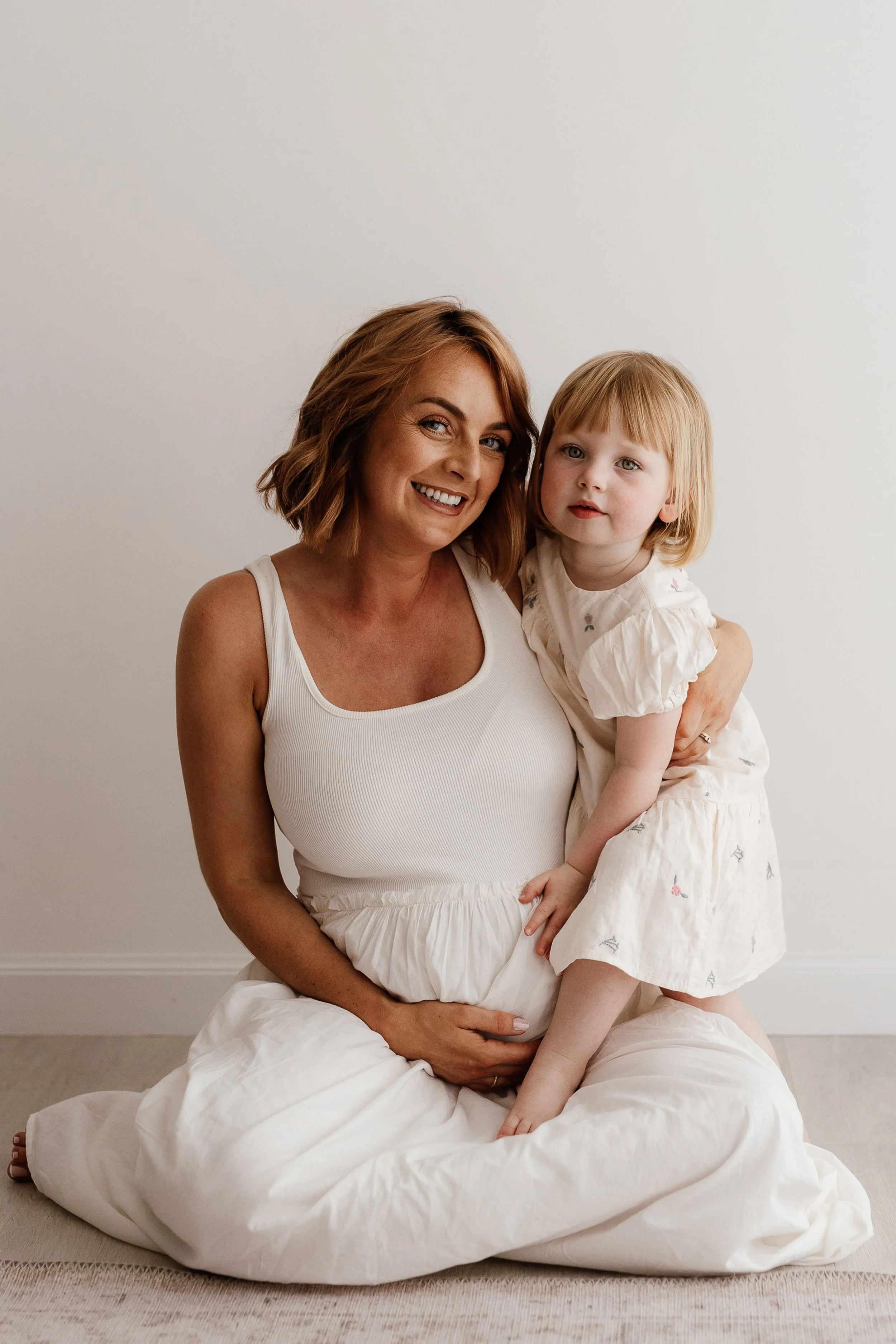 Pregnant mum sitting on the studio floor in a white dress with her young daughter cuddled on her lap.