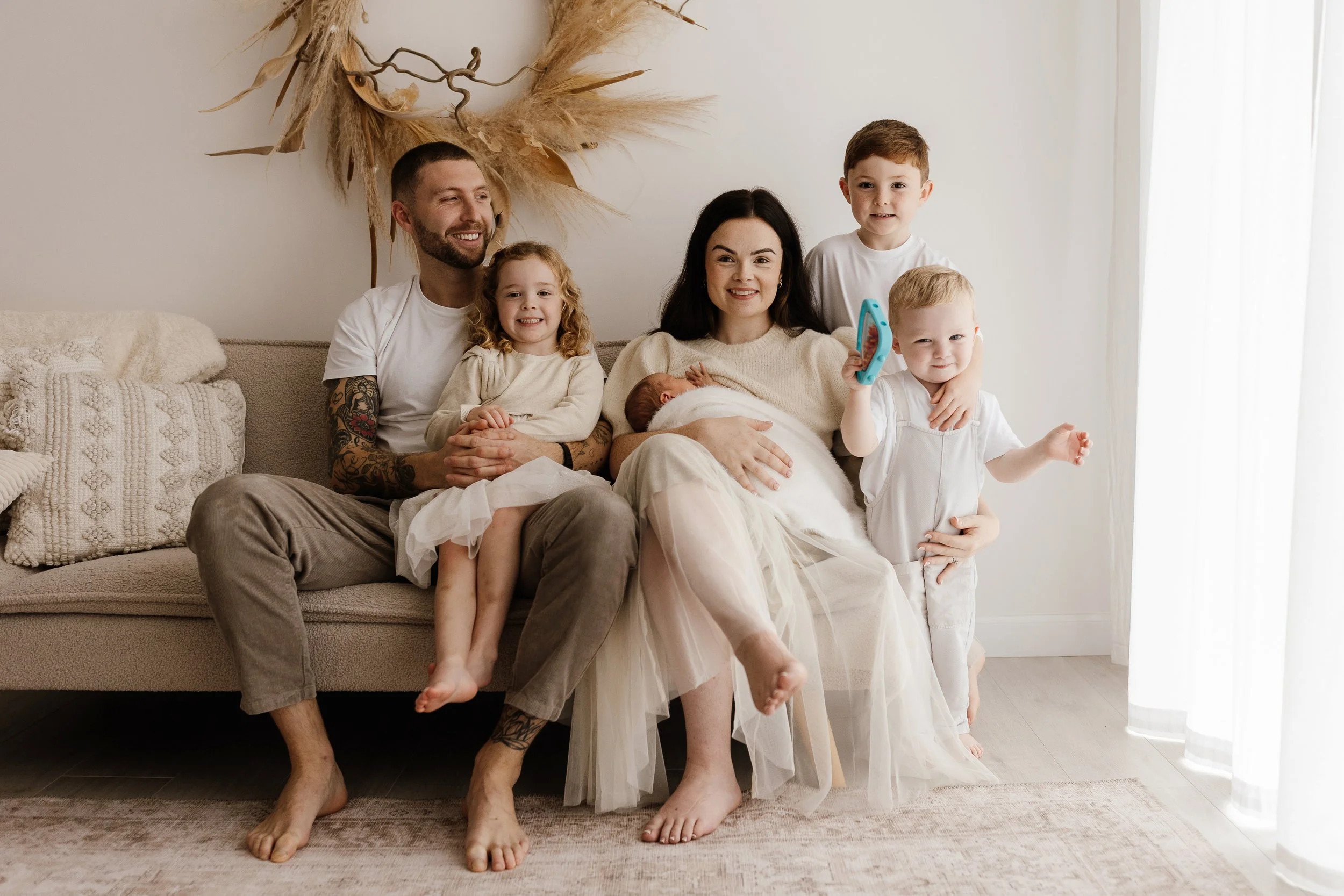  Family of five seated together on a sofa at home, mum holding a newborn while two young children and dad sit close, relaxed and smiling.