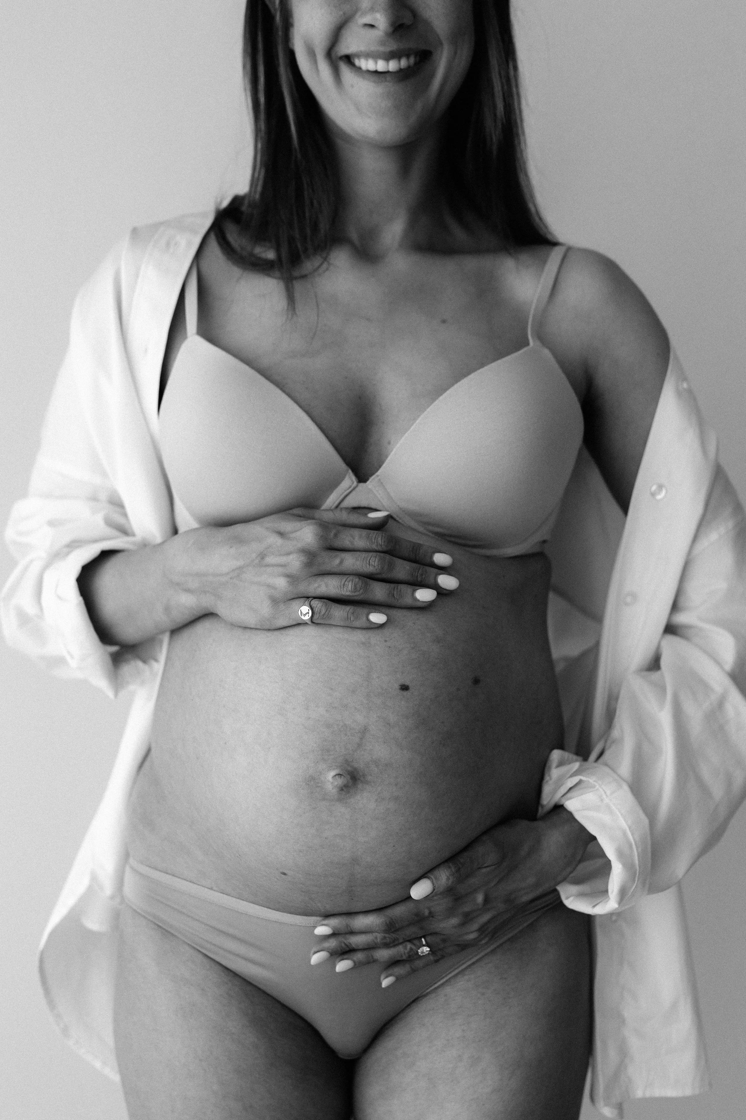 Black and white portrait of a smiling pregnant woman in underwear and an open white shirt, holding her bump.