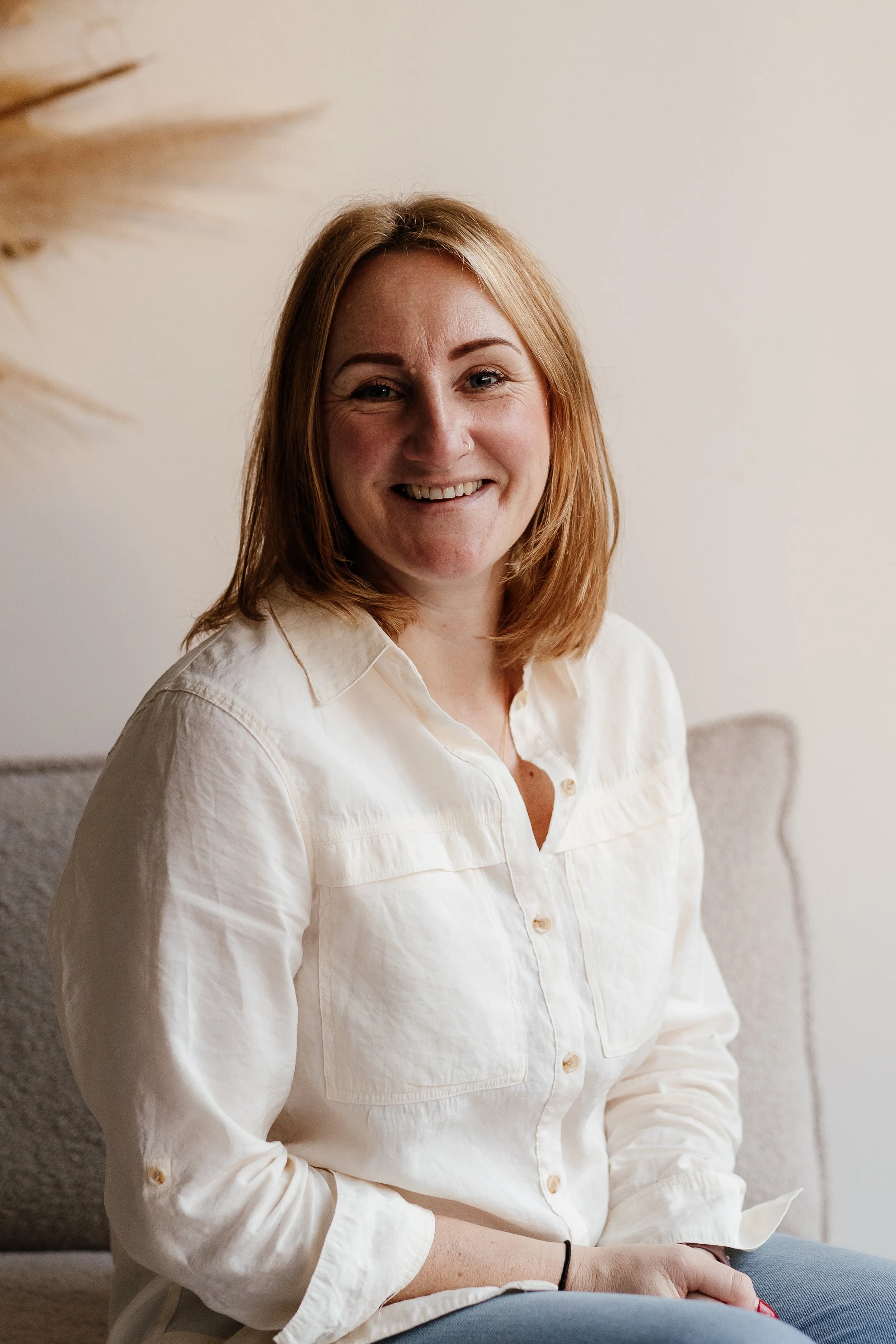 A woman with red hair smiling, wearing a cream-colored shirt, sitting indoors on a sofa with a neutral background.