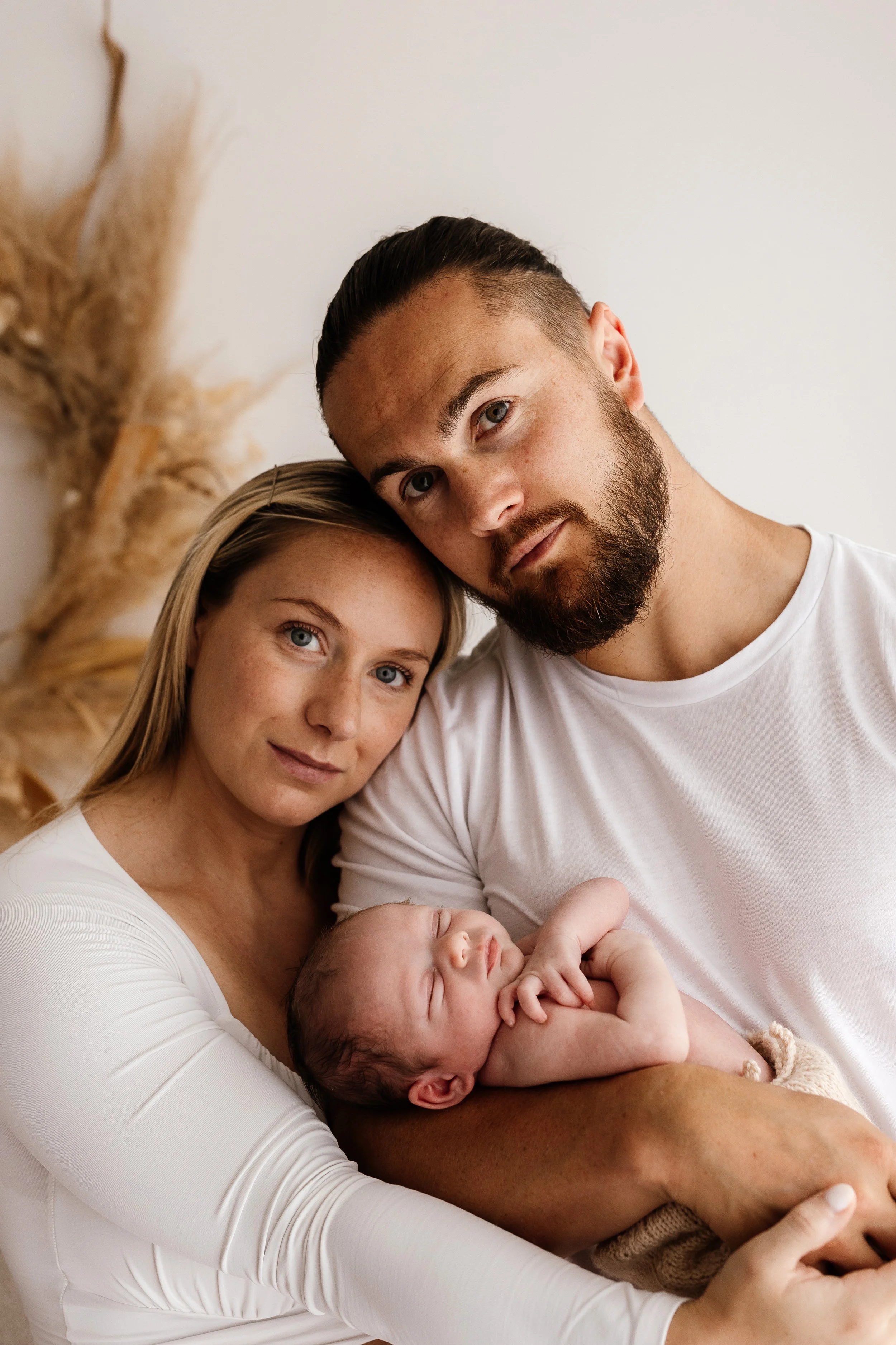 New parents holding their newborn close in soft studio light, calm family cuddle with baby sleeping in their arms.