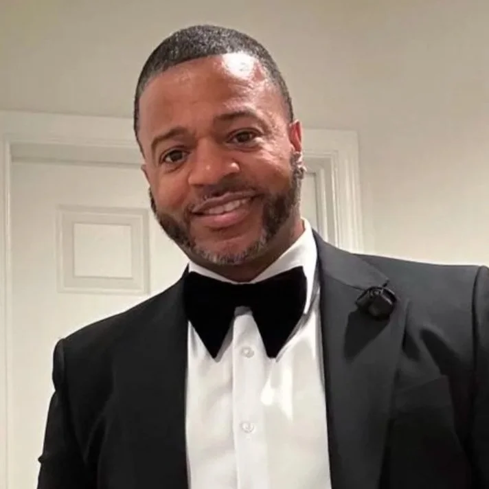 Man in a black tuxedo with a bow tie, smiling, taking a selfie indoors.