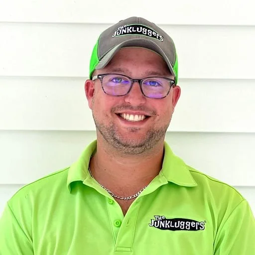 A man smiling, wearing glasses, a green polo shirt, a gray and green cap with 'The Junkluggers' logo, and a chain necklace, standing against a white wall.