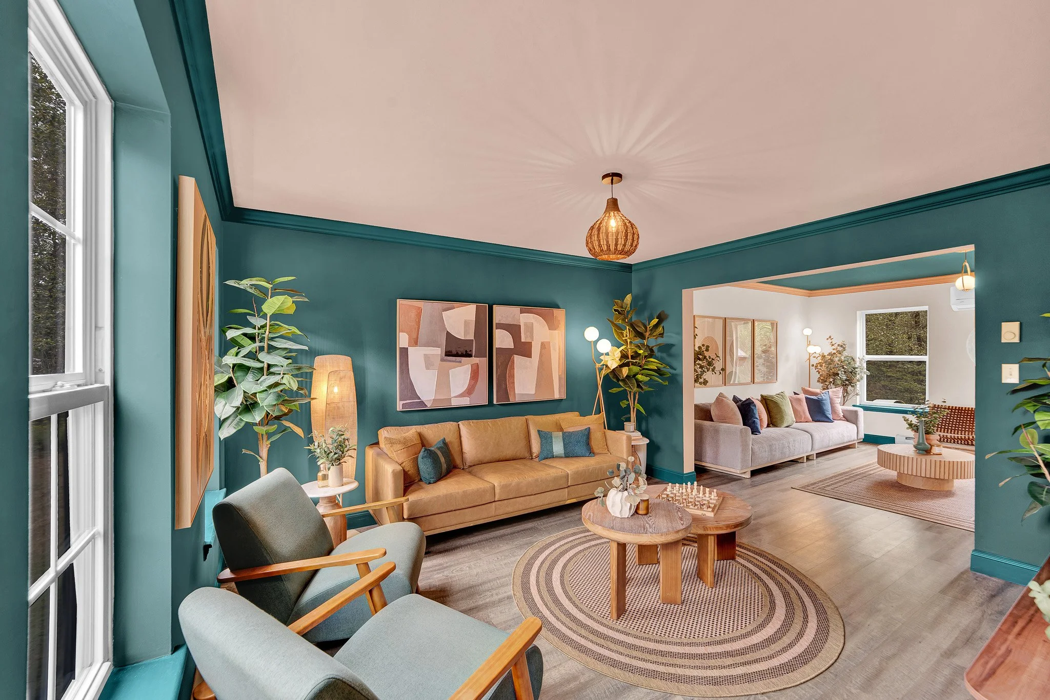 Living room with teal walls, beige and gray sofas, wooden coffee tables, large windows, plants, and modern artwork.