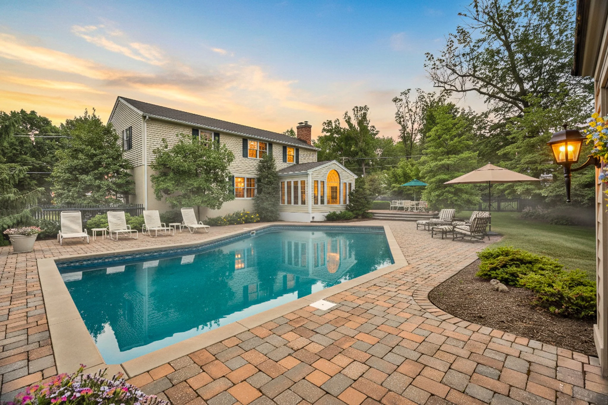 A backyard with a swimming pool, patio furniture with lounge chairs, tables with umbrellas, and a two-story house with a sunroom at sunset.