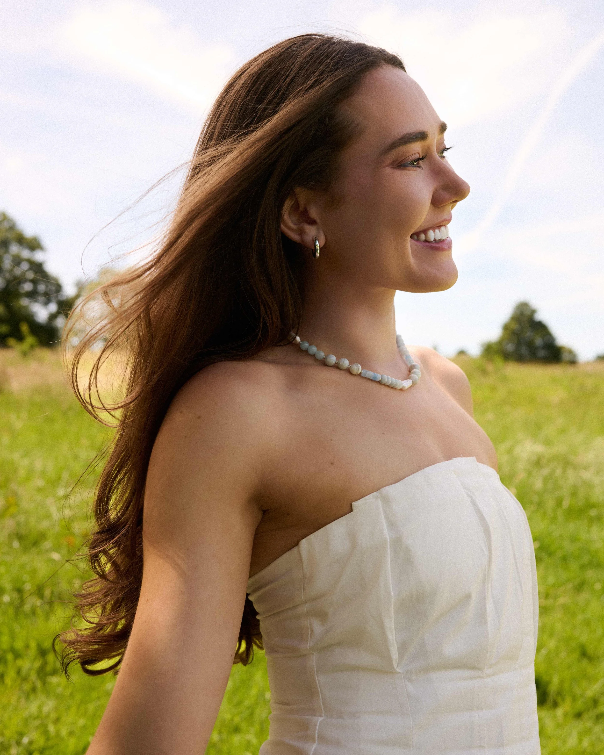 Female model in a white dress at a park, smiling and wearing a light blue beaded necklace