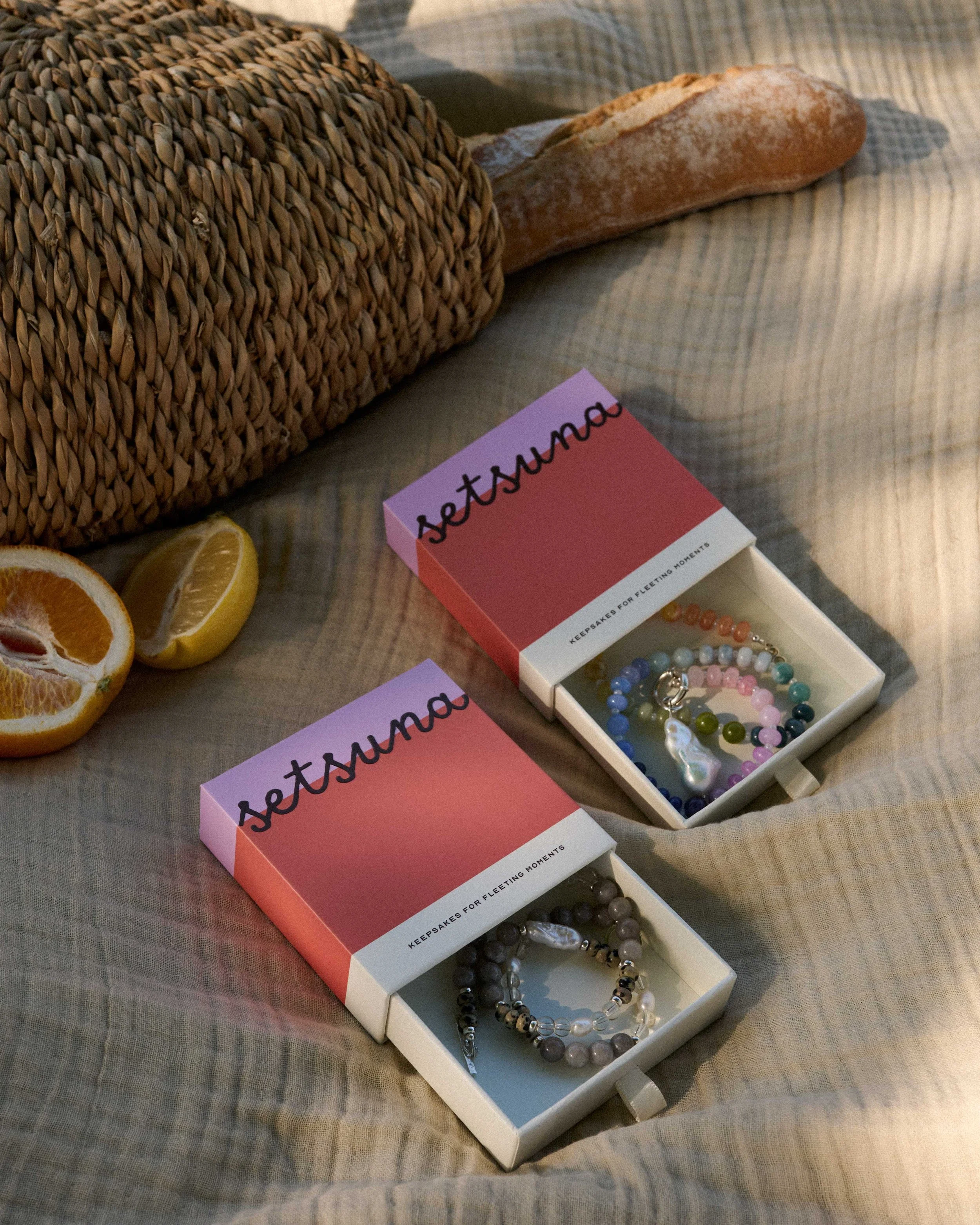 Two jewellery boxes partly open with beaded necklaces inside them, on a picnic blanket, placed next to a picnic basket