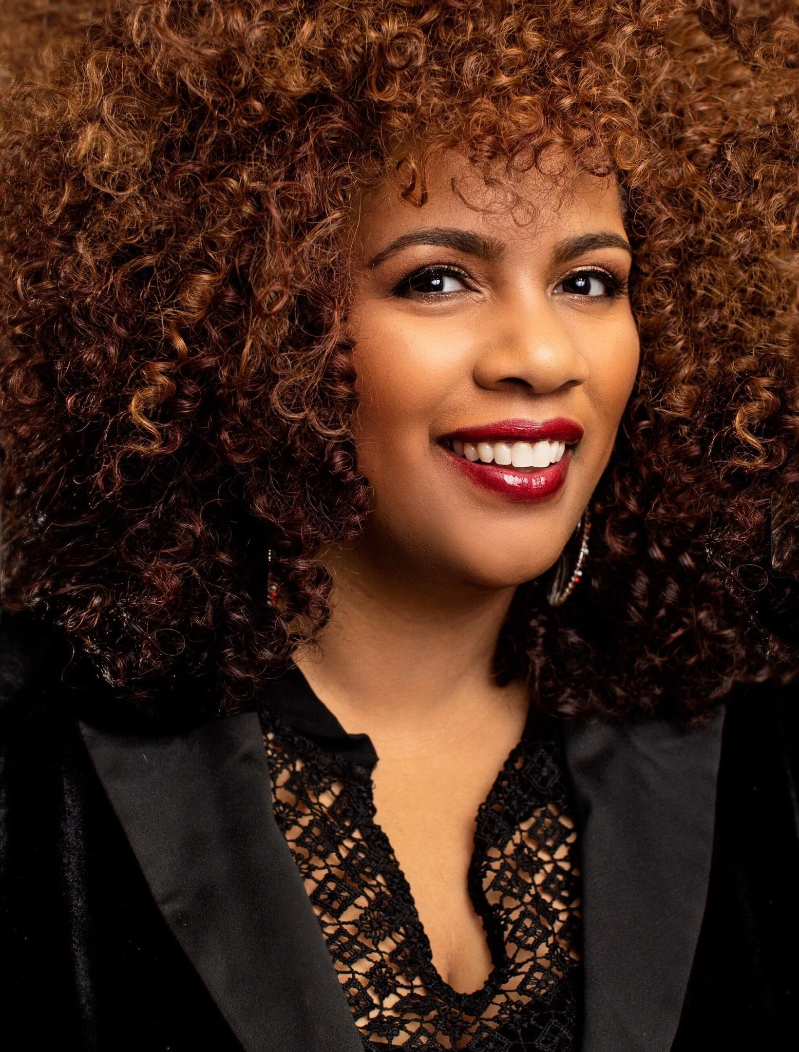 Close-up of Jetta Jewel with voluminous, curly auburn hair, wearing makeup with dark eyeliner, mascara, eyeshadow, and red lipstick, smiling with exposed teeth, dressed in a black lace top and blazer.