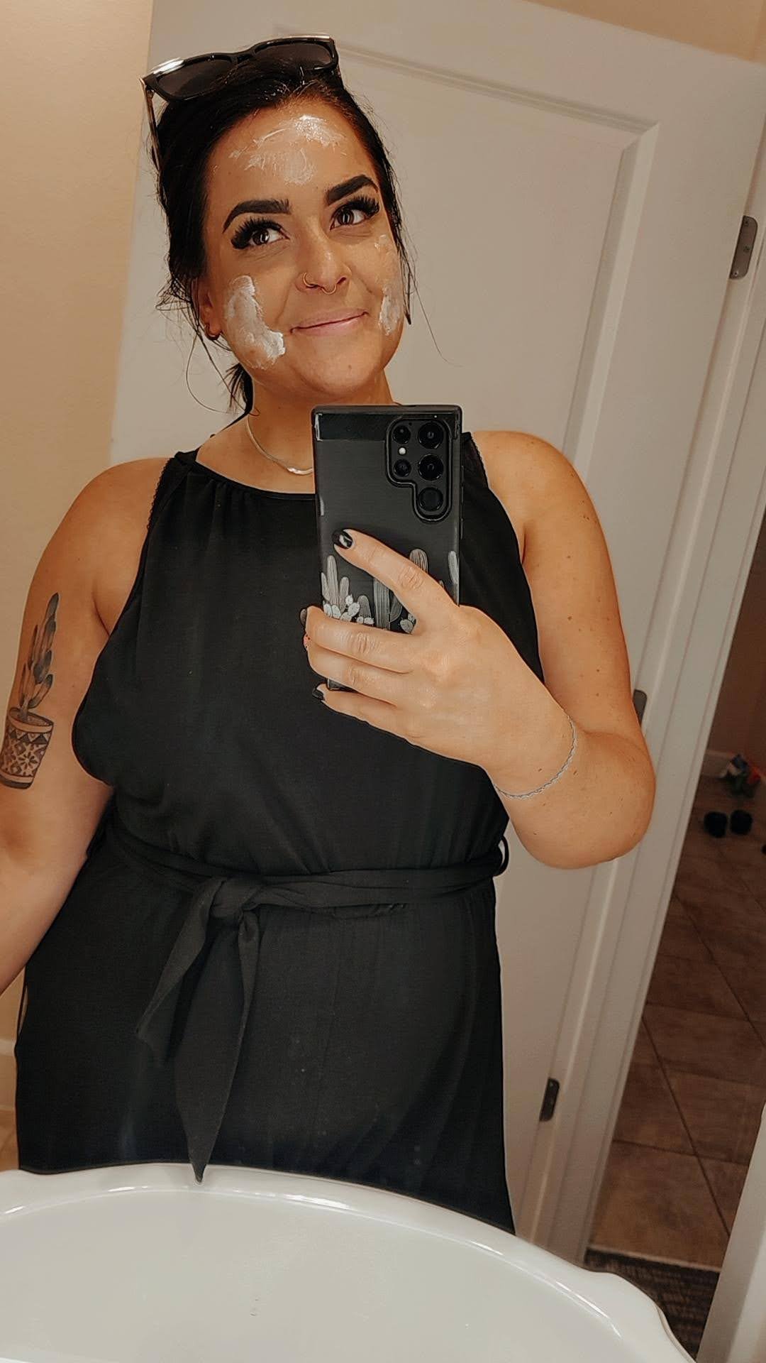 Woman taking a selfie in a bathroom mirror, wearing a black sleeveless top, with a tattoo on her left arm, and sunglasses on her head. She has soap or cream on her face.