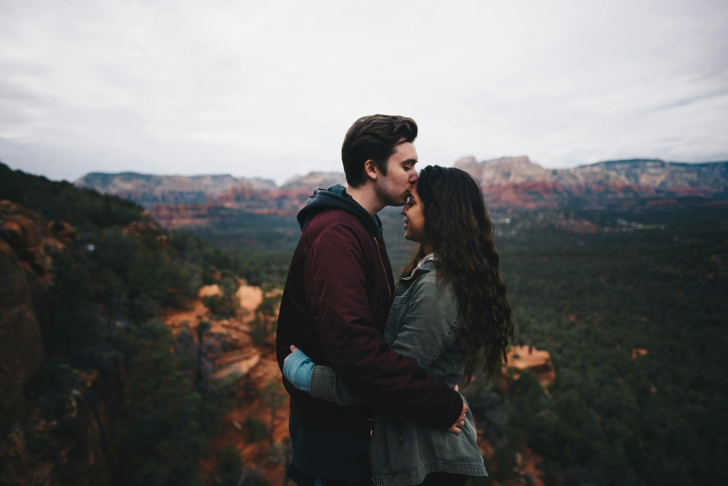 A young couple embraces and shares a kiss in front of a scenic canyon landscape with layered rock formations and a cloudy sky.