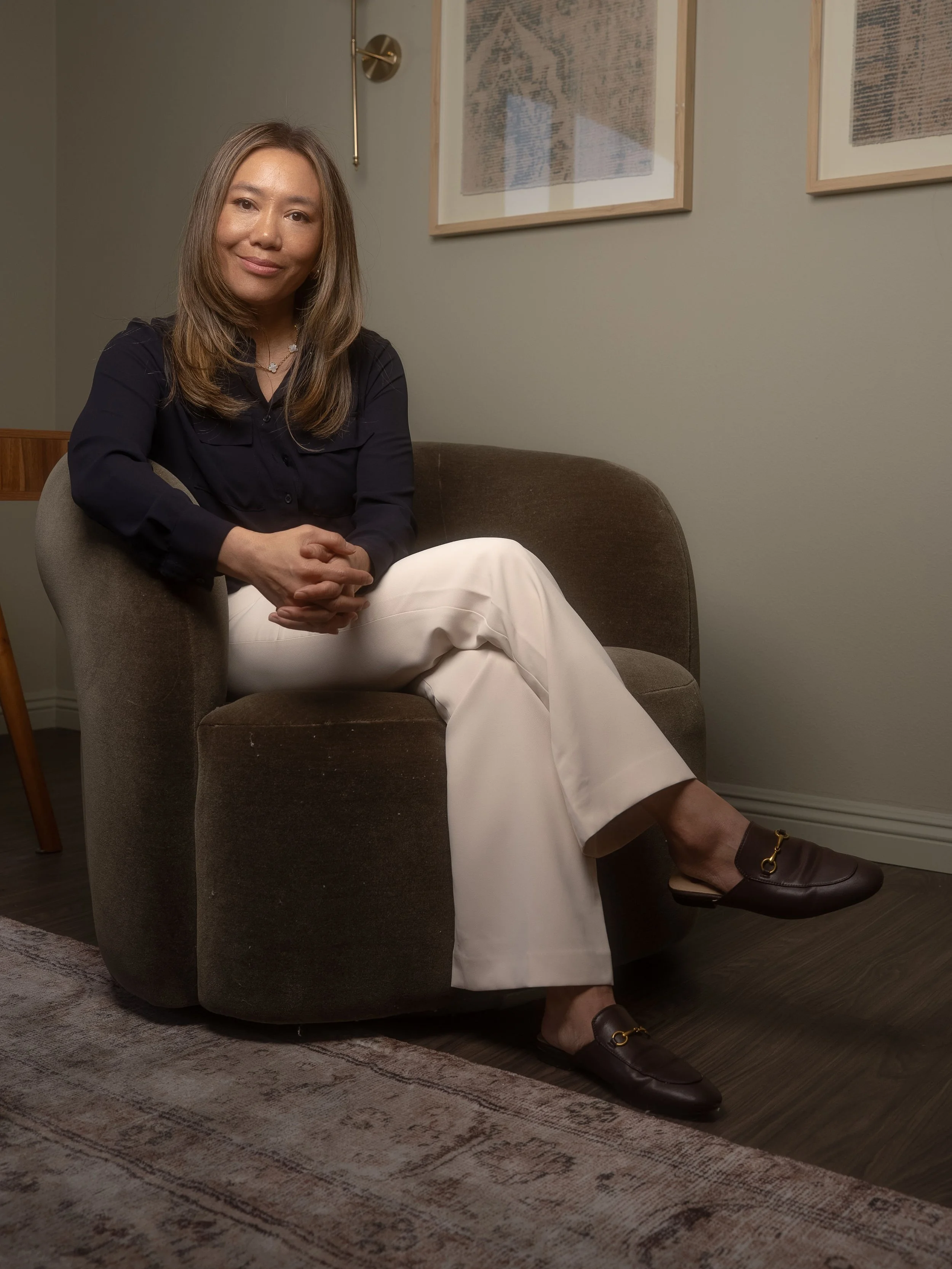 A woman sitting on a brown chair with crossed legs, wearing a black blouse, white pants, and brown loafers, indoors with framed art on the wall behind her.