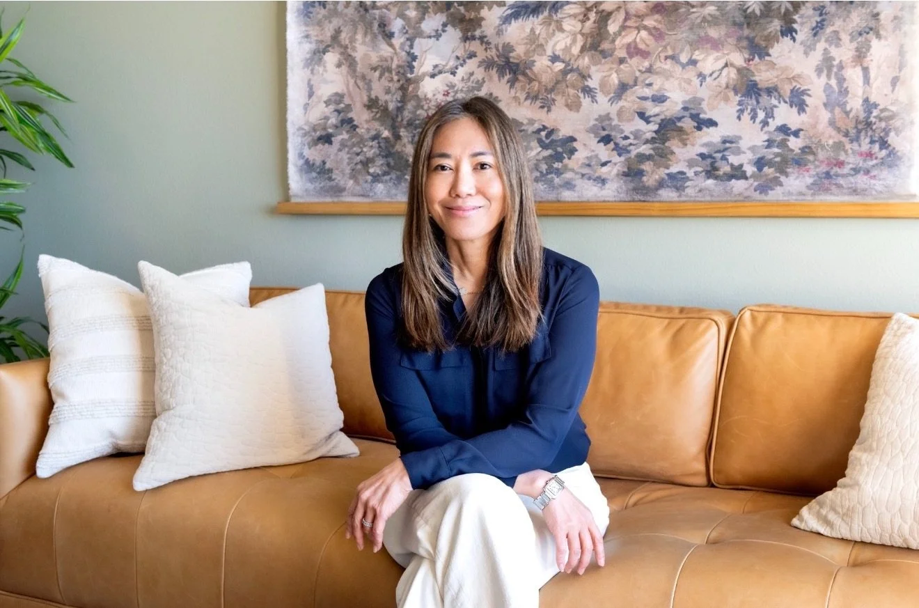 A woman with long brown hair sitting on a tan leather sofa with white pillows, smiling and wearing a navy blue blouse and white pants. A plant is on the left and a large framed artwork hangs on the green wall behind her.