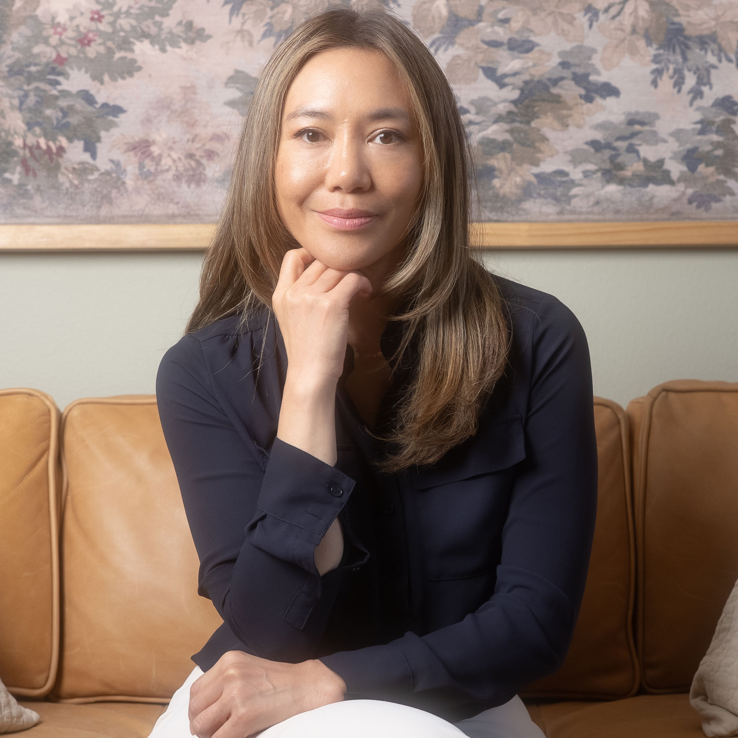 A woman with long brown hair wearing a navy blue shirt, sitting on a tan leather couch, resting her chin on her hand, smiling slightly, with a floral painting in the background.
