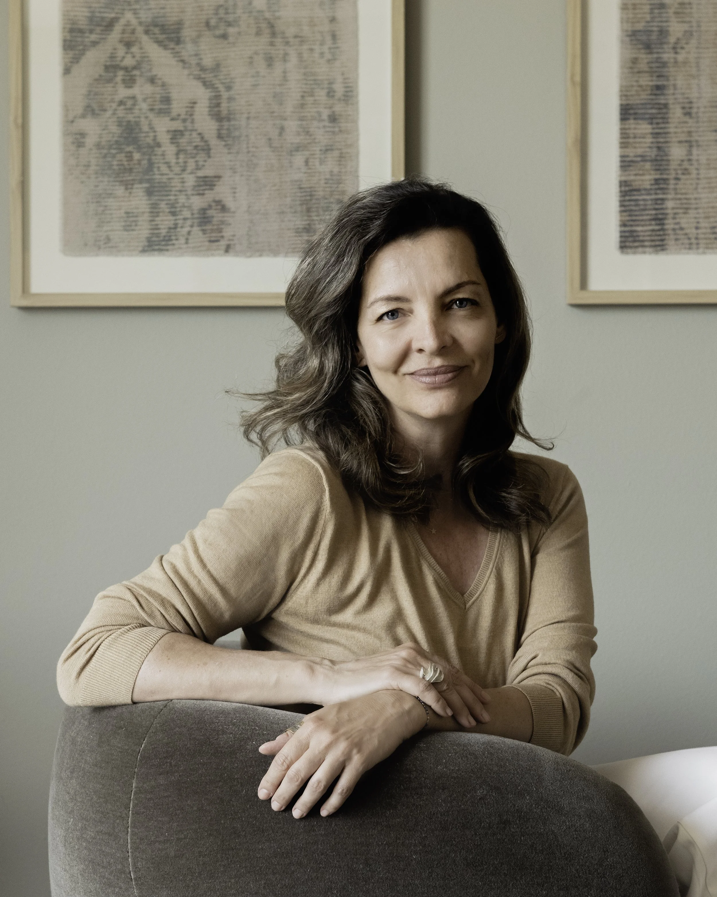 A woman with shoulder-length brown hair and a beige shirt sitting on a chair, smiling, with framed artwork on the wall behind her.