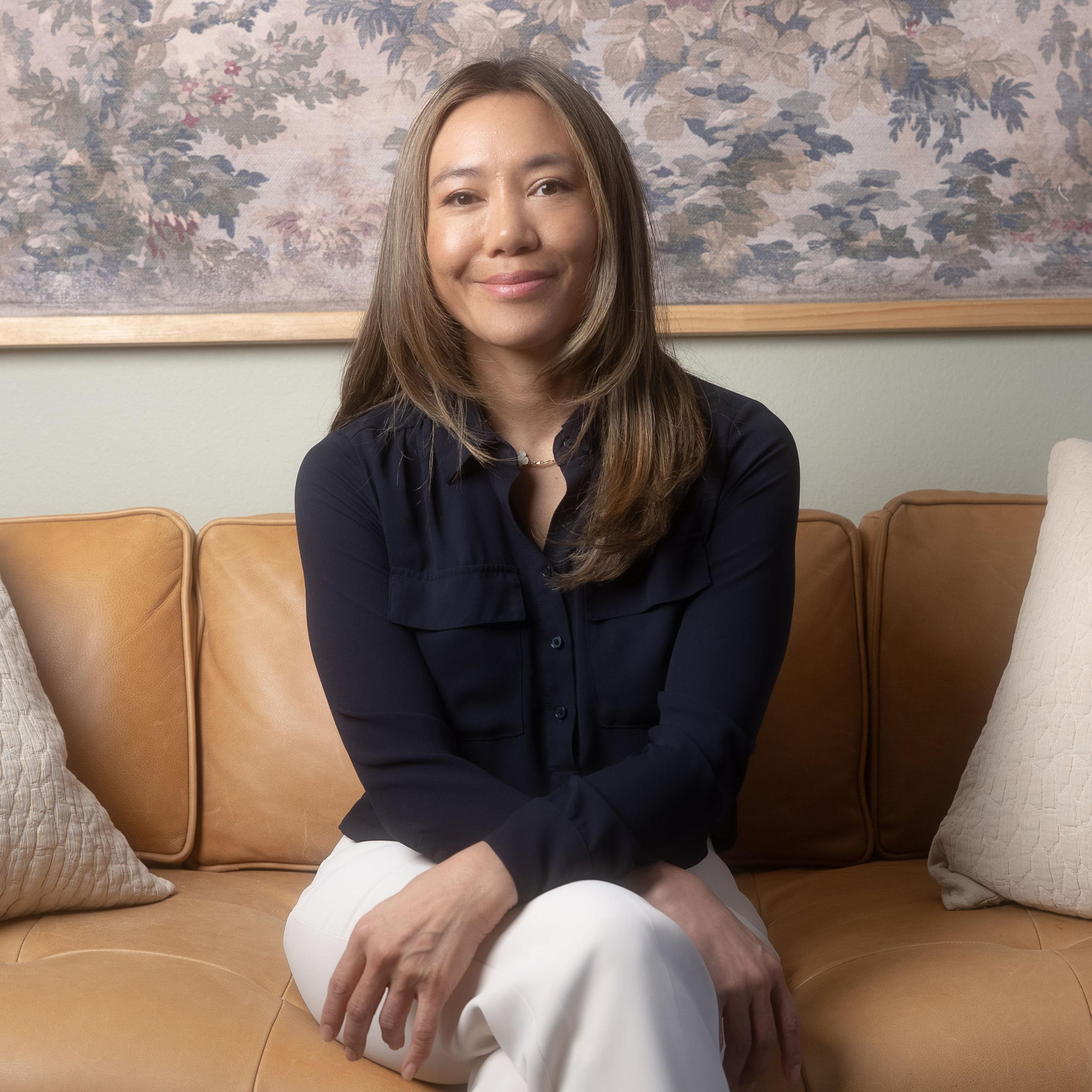 A woman with shoulder-length brown hair, wearing a black blouse and white pants, sitting on a tan leather sofa with cream and beige pillows, in front of a floral wall art.