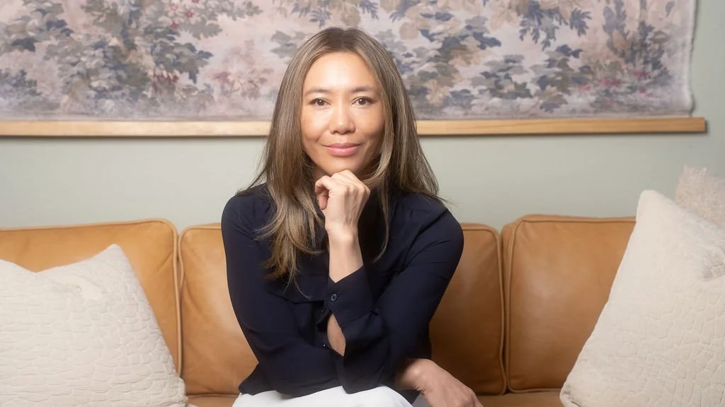 A woman with long hair sitting on a tan leather couch, resting her chin on her hand, in front of a floral tapestry background.