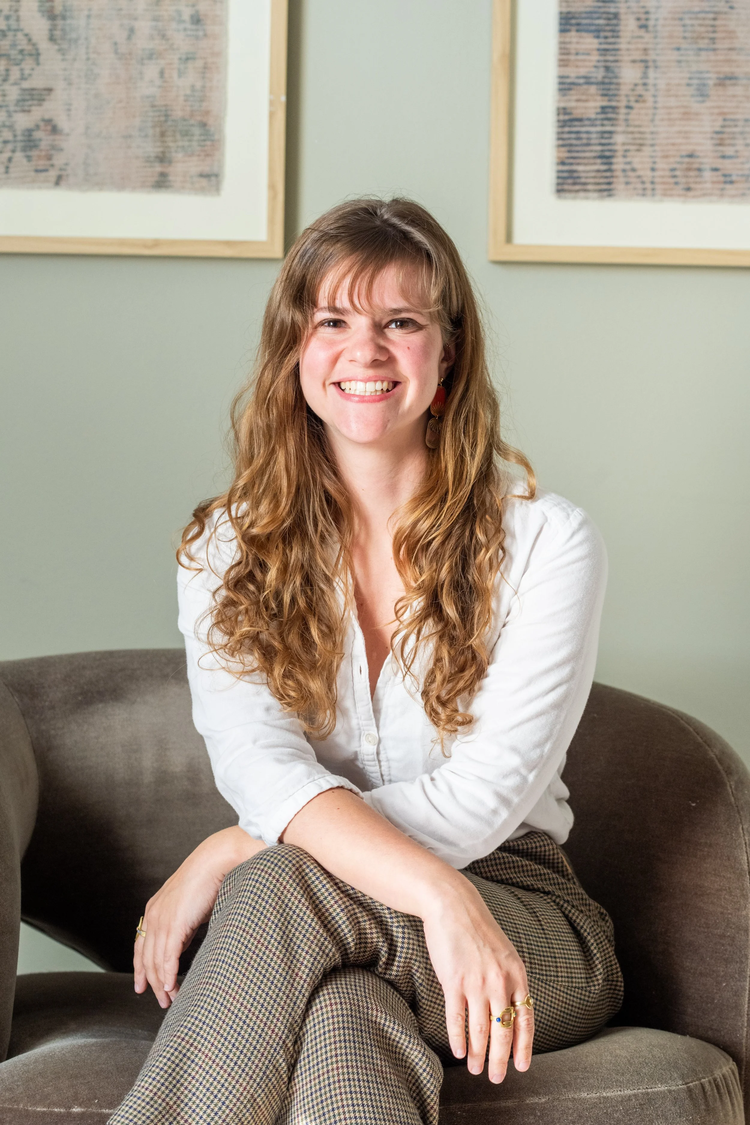 A woman with long curly hair and a white shirt sitting on a brown sofa, smiling with art on the wall behind her.