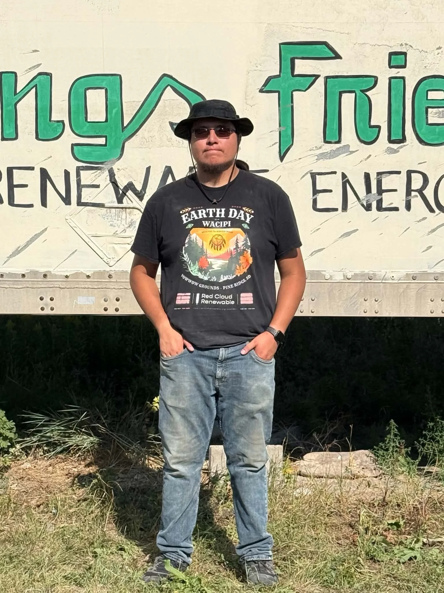 A person standing outdoors in front of a large sign that reads 'Energy', 'Renewable', and 'Energy' with various partially visible words and symbols. The person is wearing a black wide-brimmed hat, sunglasses, a black T-shirt with Earth Day and environmental graphics, jeans, and sneakers.