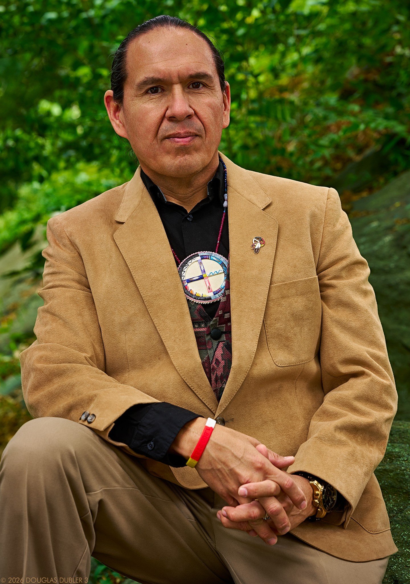 Portrait of a man with long dark hair tied in a braid, wearing a blue shirt and a dark blazer, with colorful bead necklaces and jewelry, smiling against a gray background.