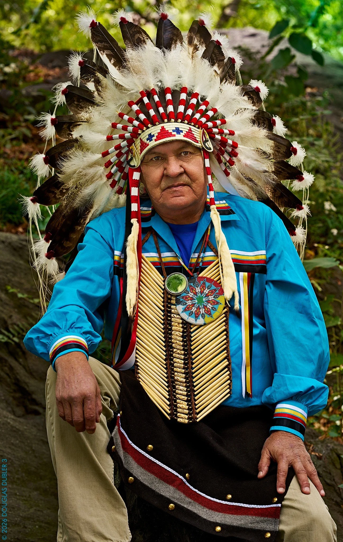 Man dressed in traditional Native American headdress and clothing standing outdoors with mountains in the background.
