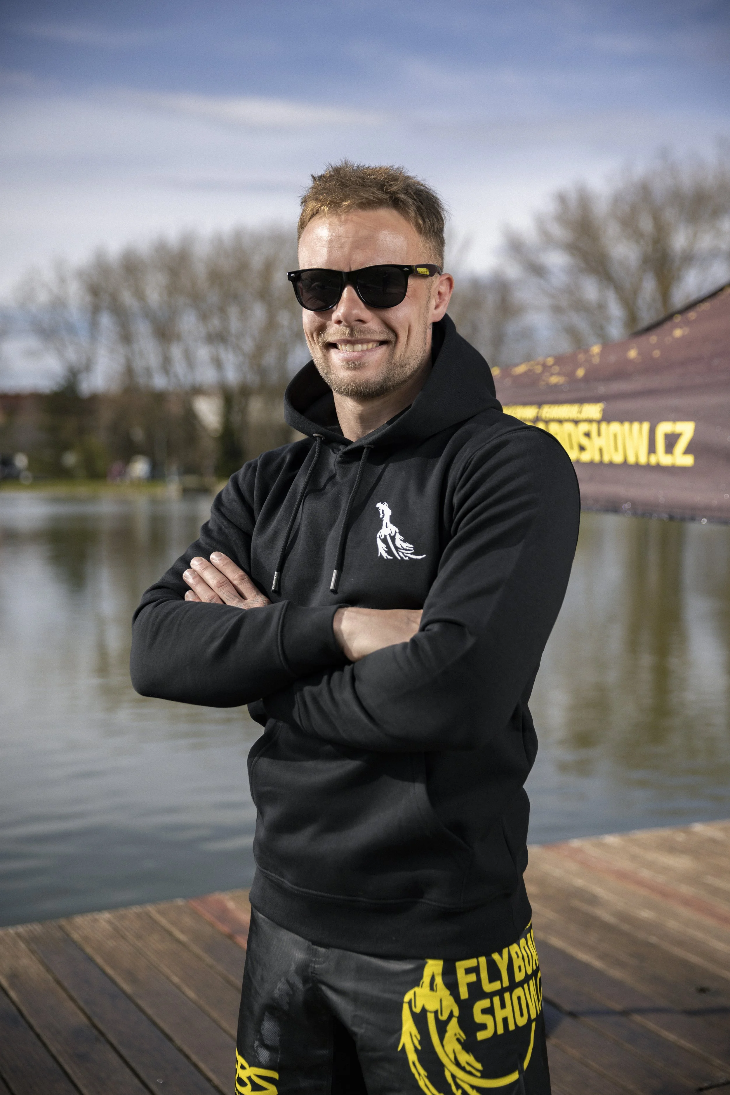 Man wearing black hoodie and shorts with yellow logo, sunglasses, smiling with arms crossed by a lake during daytime.