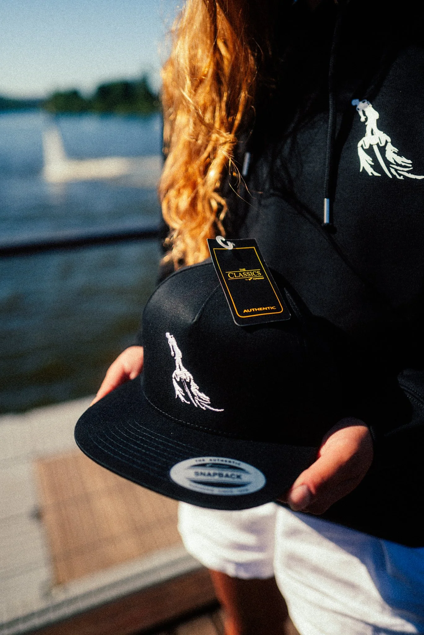 Person holding a black cap with a white embroidered lighthouse design, near water with a sailboat in the background, wearing a black jacket with a logo, and white shorts.