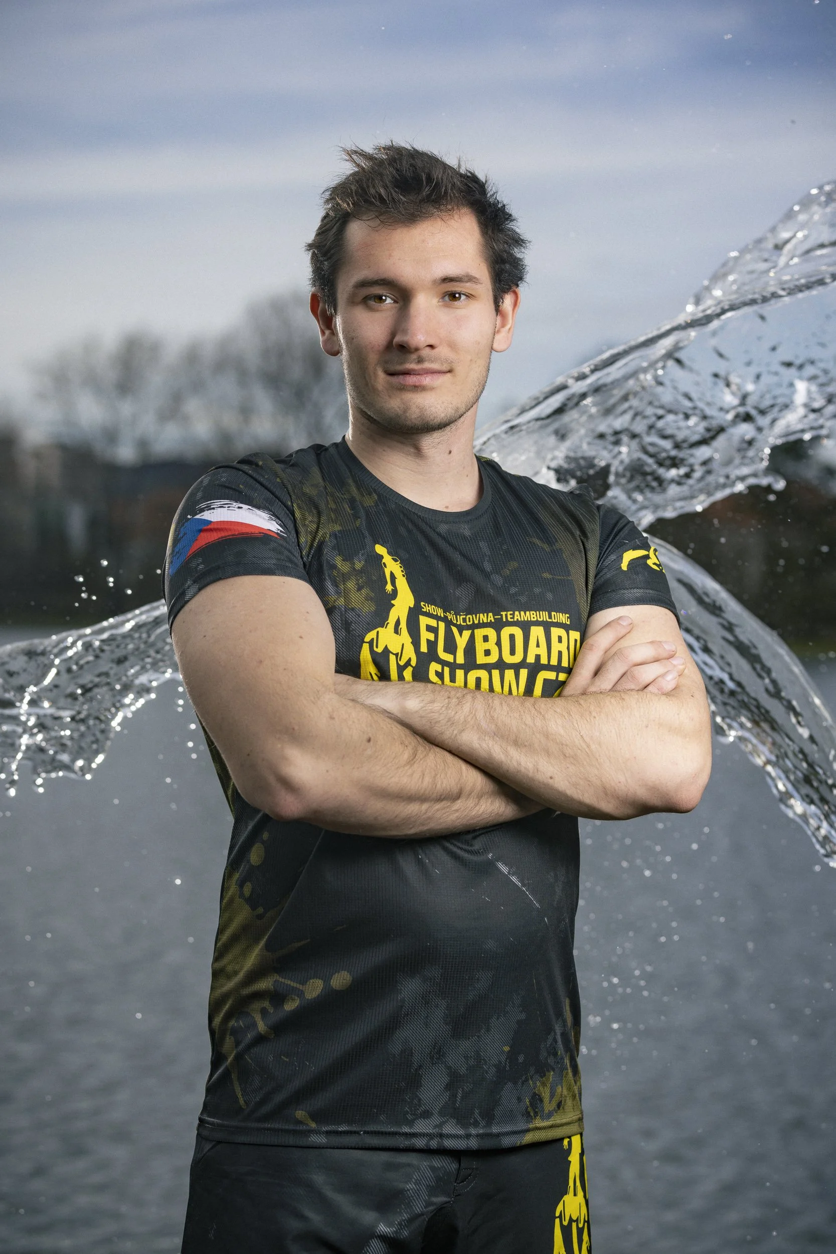 A young man with arms crossed standing outdoors near a body of water, with a splash of water in the background, wearing a black sports shirt with yellow, red, blue, and white accents and the words 'Flyboard Show' printed on it.