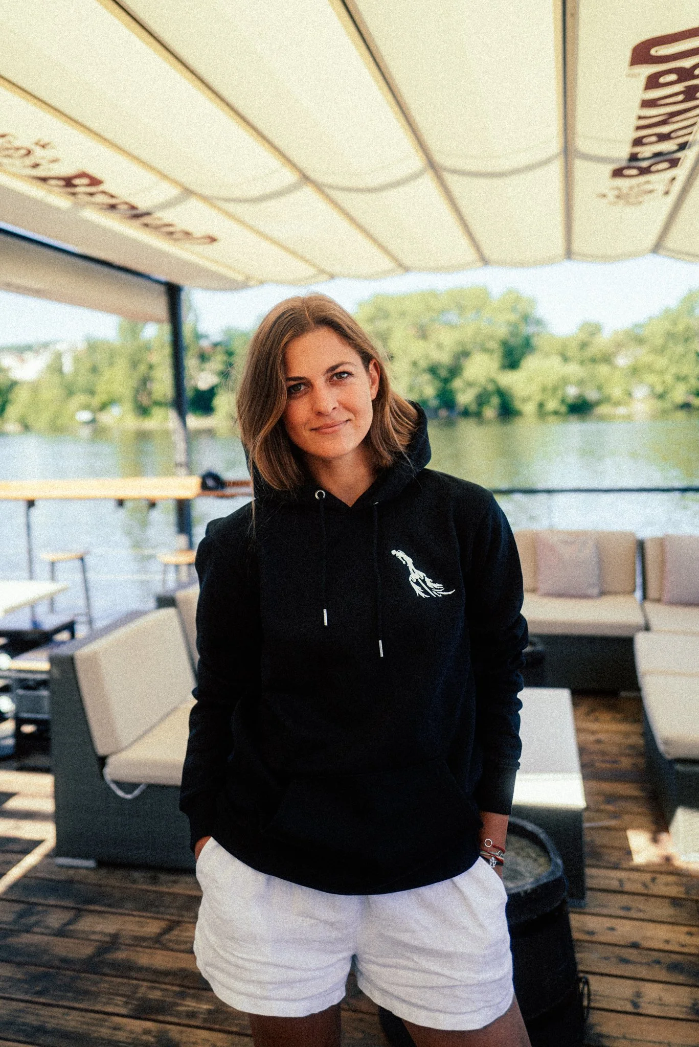 A woman with shoulder-length brown hair wearing a black hoodie and white shorts standing on a deck by the water, with trees and a cloudy sky in the background.
