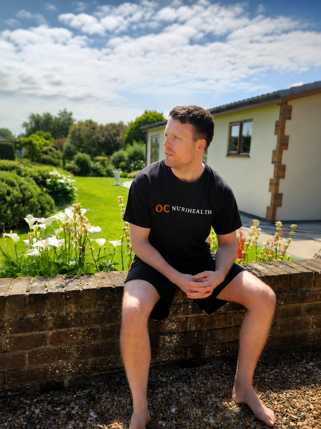 A man sitting on a brick wall in a garden, wearing a black T-shirt and shorts, with a house and greenery in the background on a sunny day.