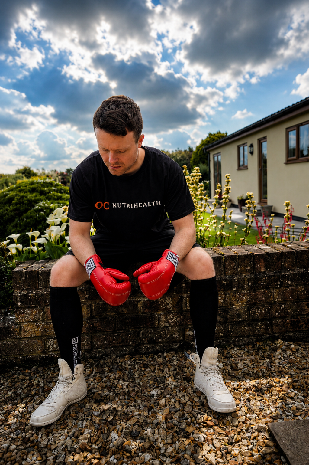 Man sitting on a brick wall wearing red boxing gloves, black sportswear, and white sneakers, in a garden with flowers and a house in the background under a partly cloudy sky.