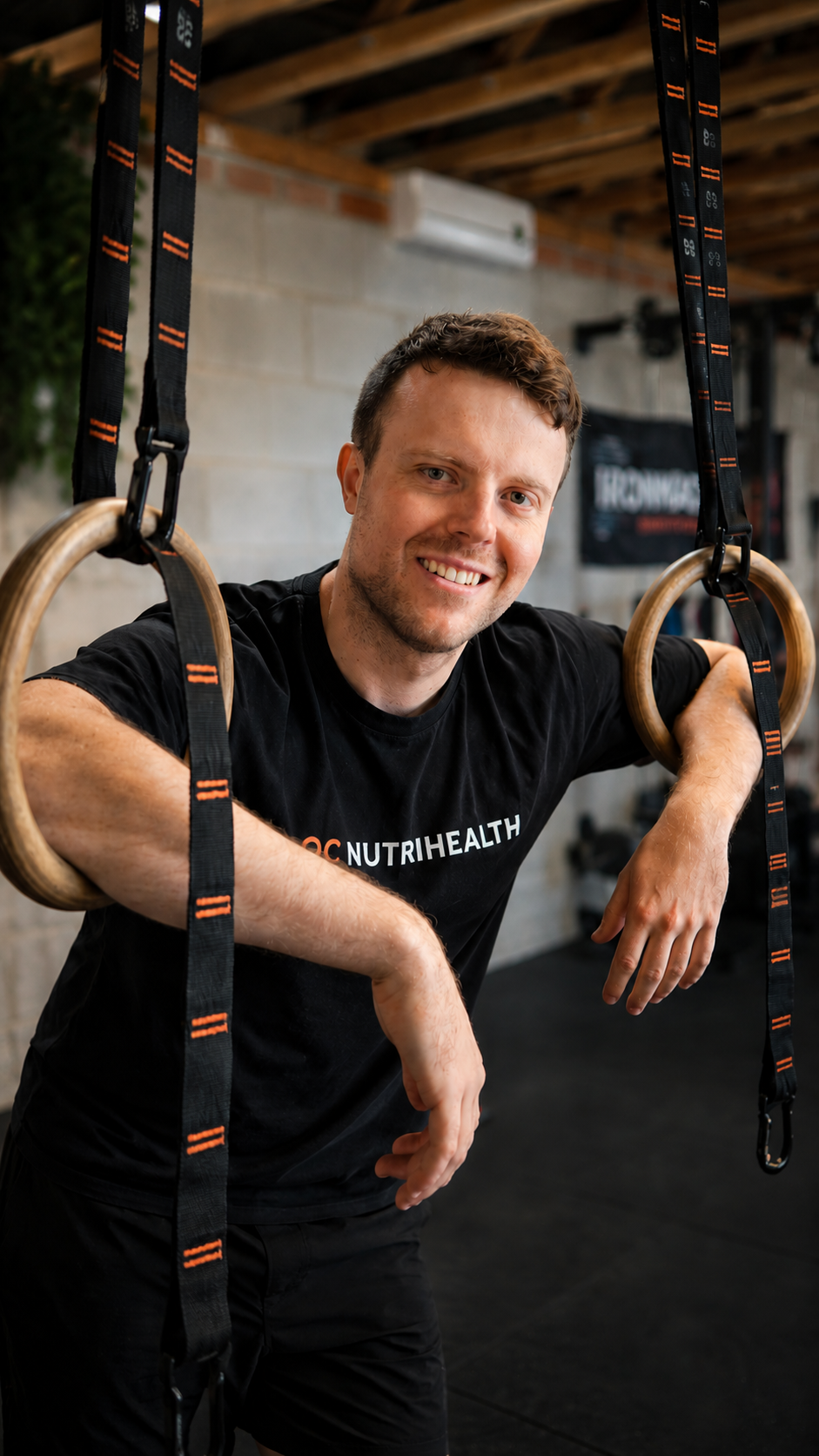 A man smiling and leaning on gymnastic rings in a fitness gym, wearing a black T-shirt with 'NUTRIHEALTH' printed on it.