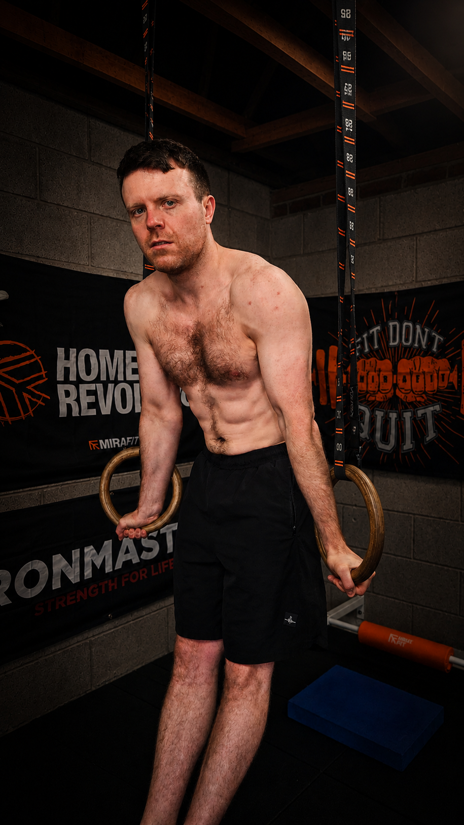 A shirtless man with short dark hair and light skin performs a workout with gymnastic rings in a gym, surrounded by motivational banners and workout equipment.