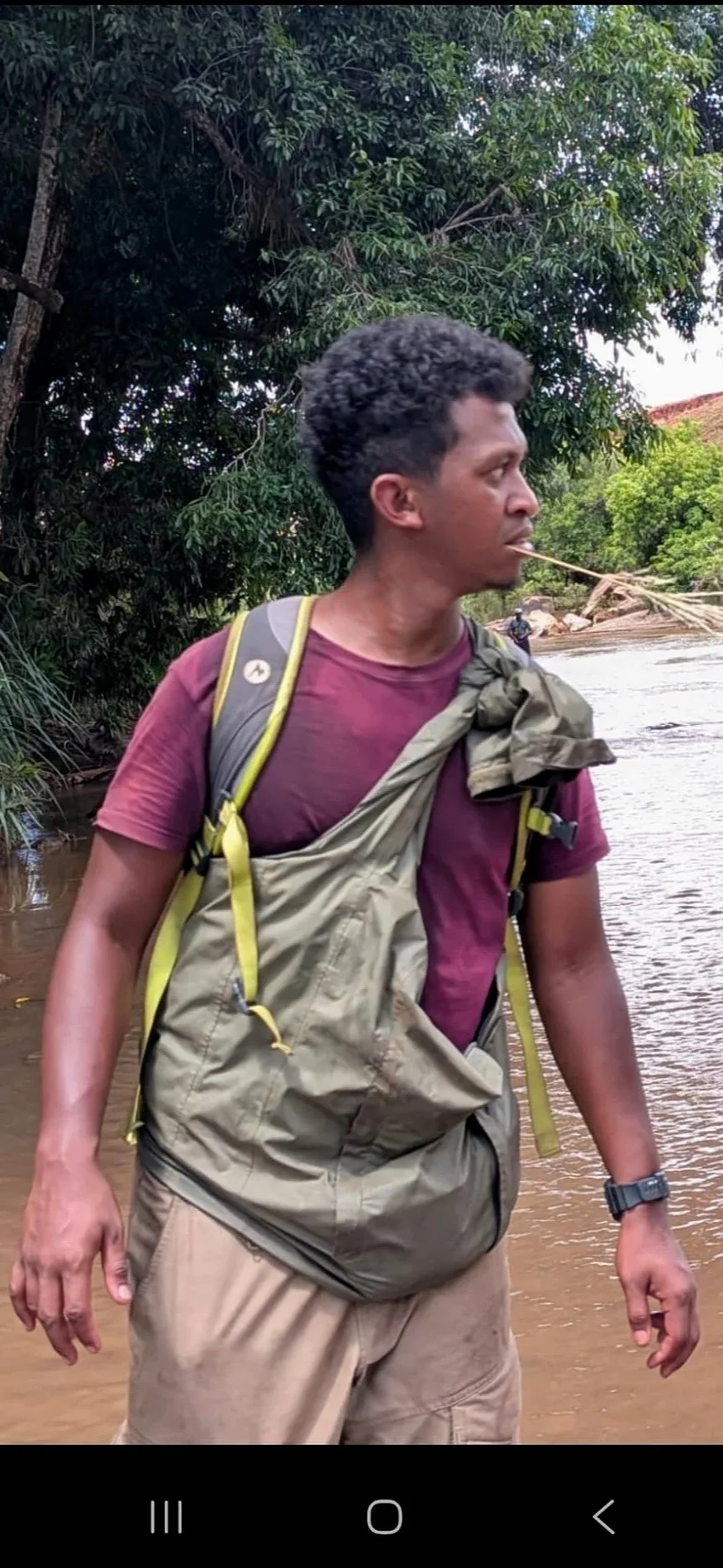 Person with a backpack, wearing a mask and a cap, standing near a riverbank in a forested area.