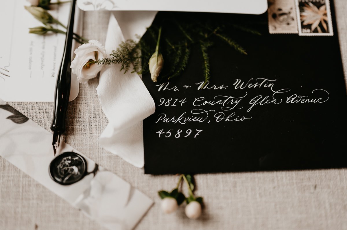 Photo showcasing wedding day details in a stylish flat lay, including the invitation suite, place cards, vintage stamps, delicate flowers, and a decorative ring box, creating a chic and cohesive look.