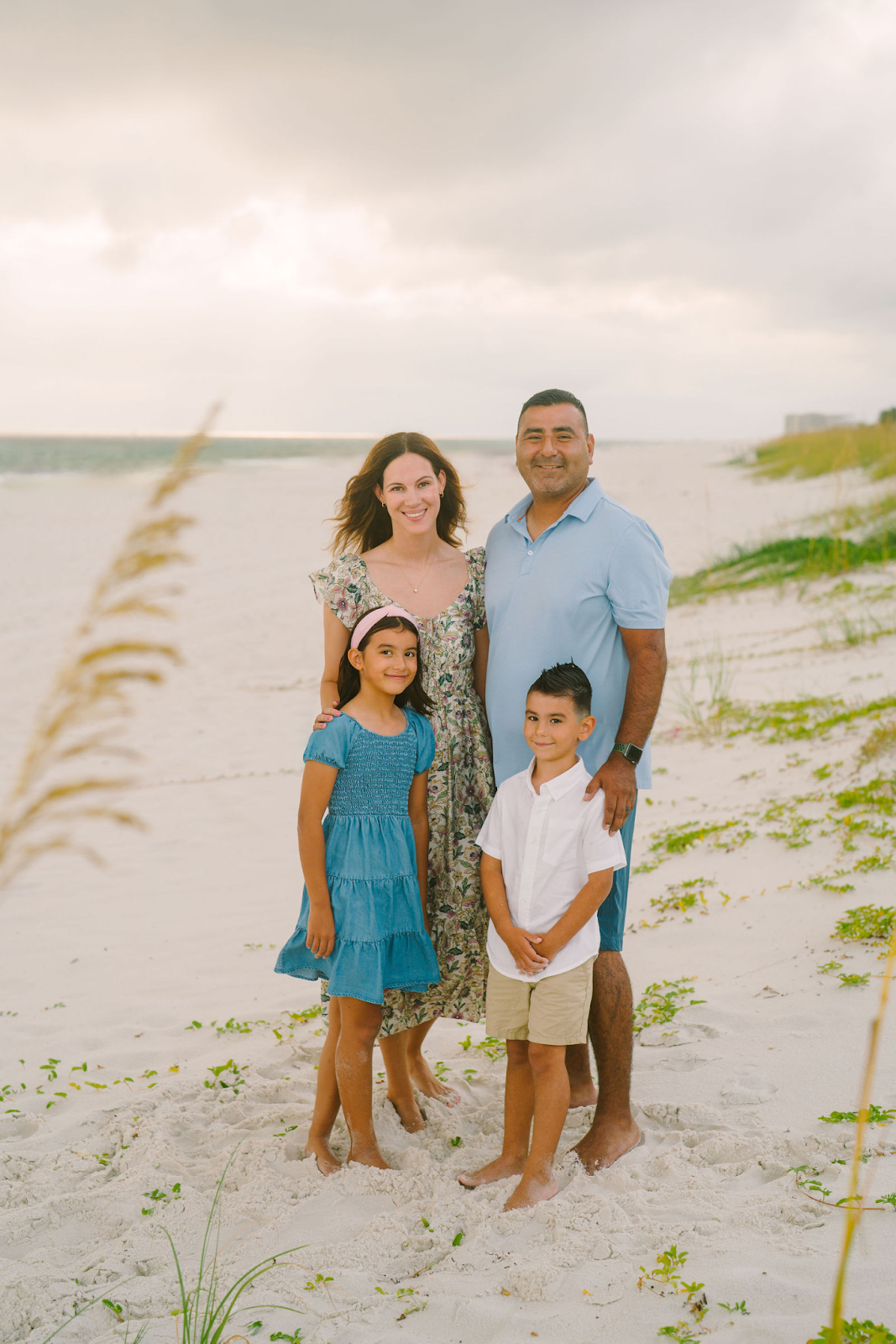 A family of four posing barefoot on a sandy beach near beach grass and dunes under a cloudy sky.