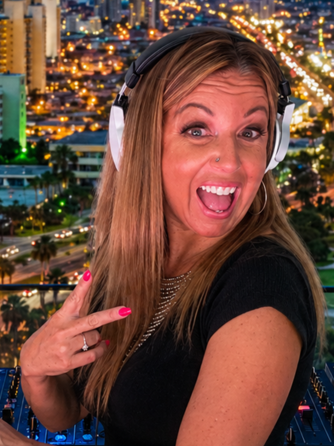 A woman with long blonde hair, wearing a black shirt, headphones, and showing a peace sign, smiling excitedly against an urban cityscape at night with lights and buildings.