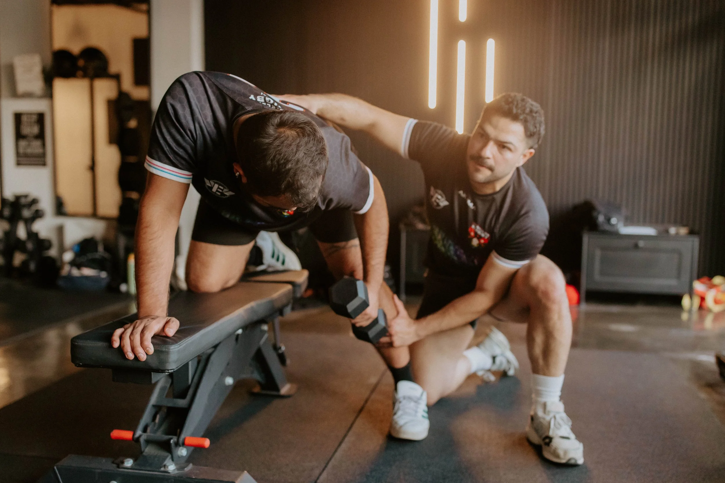 Two men in a gym, with one man helping the other do a plank exercise with dumbbells on a weight bench.
