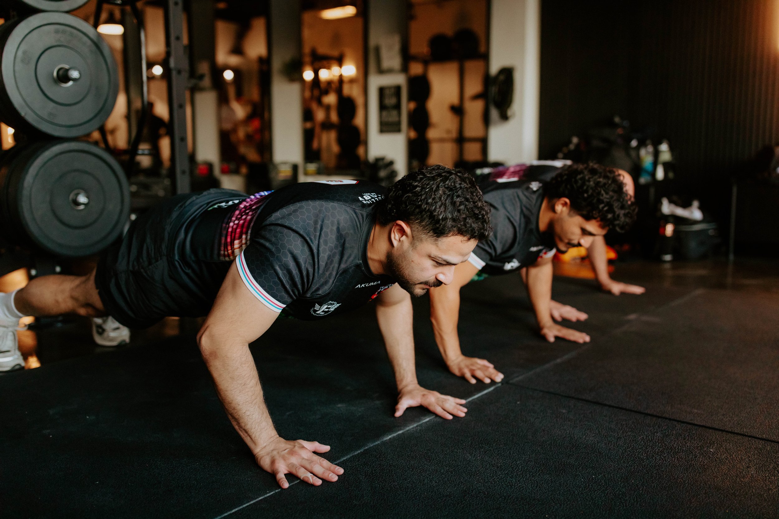 Two men in black athletic shirts with red and white trim on the sleeves doing push-ups on a black gym floor, with weights on racks in the background.