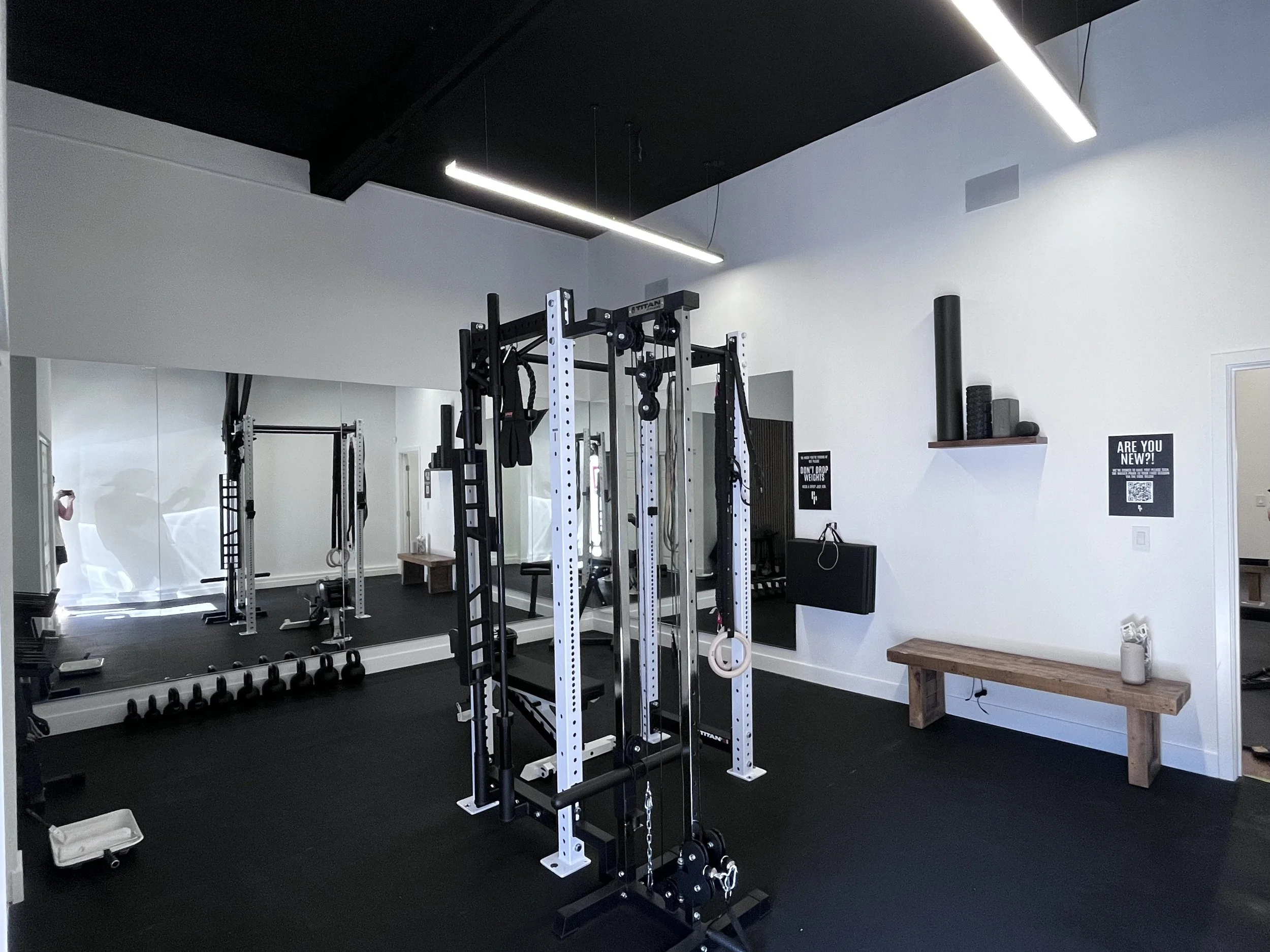 A spacious gym with black and white walls, featuring various weightlifting equipment including racks, dumbbells, and a bench. Modern lighting fixtures hang from the ceiling, and motivational signs are on the walls.