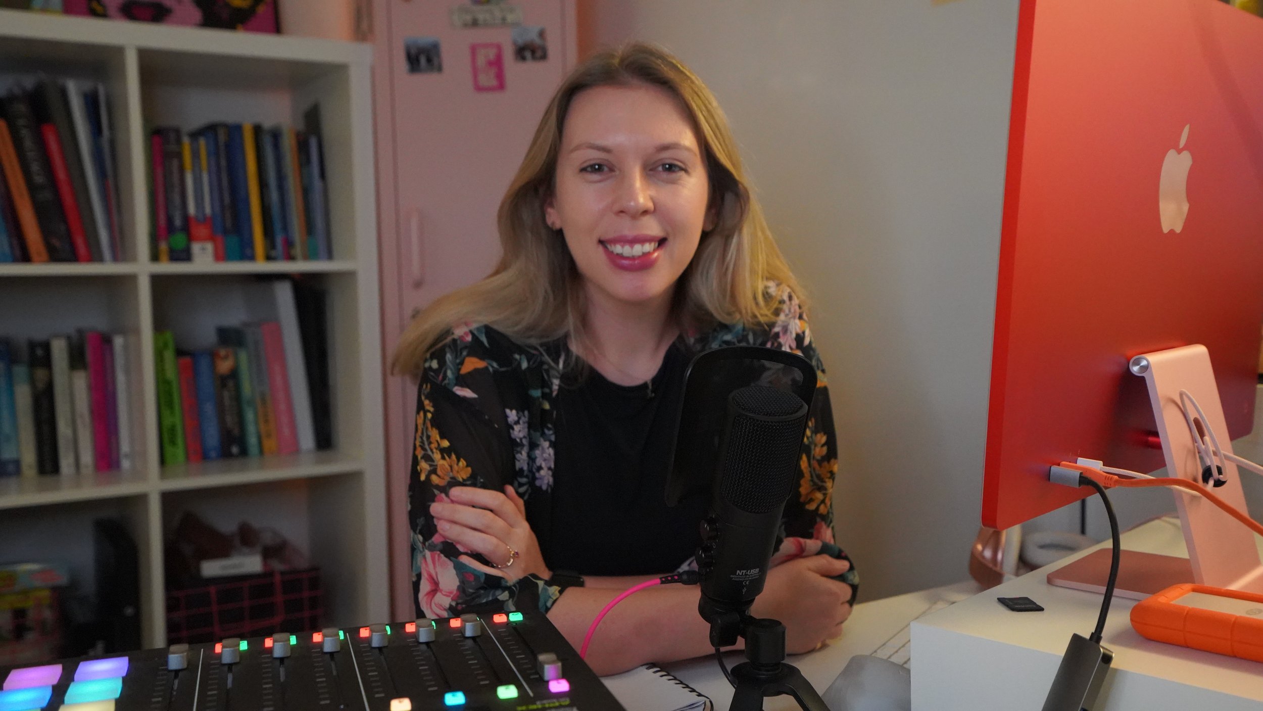 A woman with blonde hair smiling at the camera, sitting at a desk with a microphone, a mixing console, and a pink iPad in front of a red computer monitor. A bookshelf with colorful books is in the background.