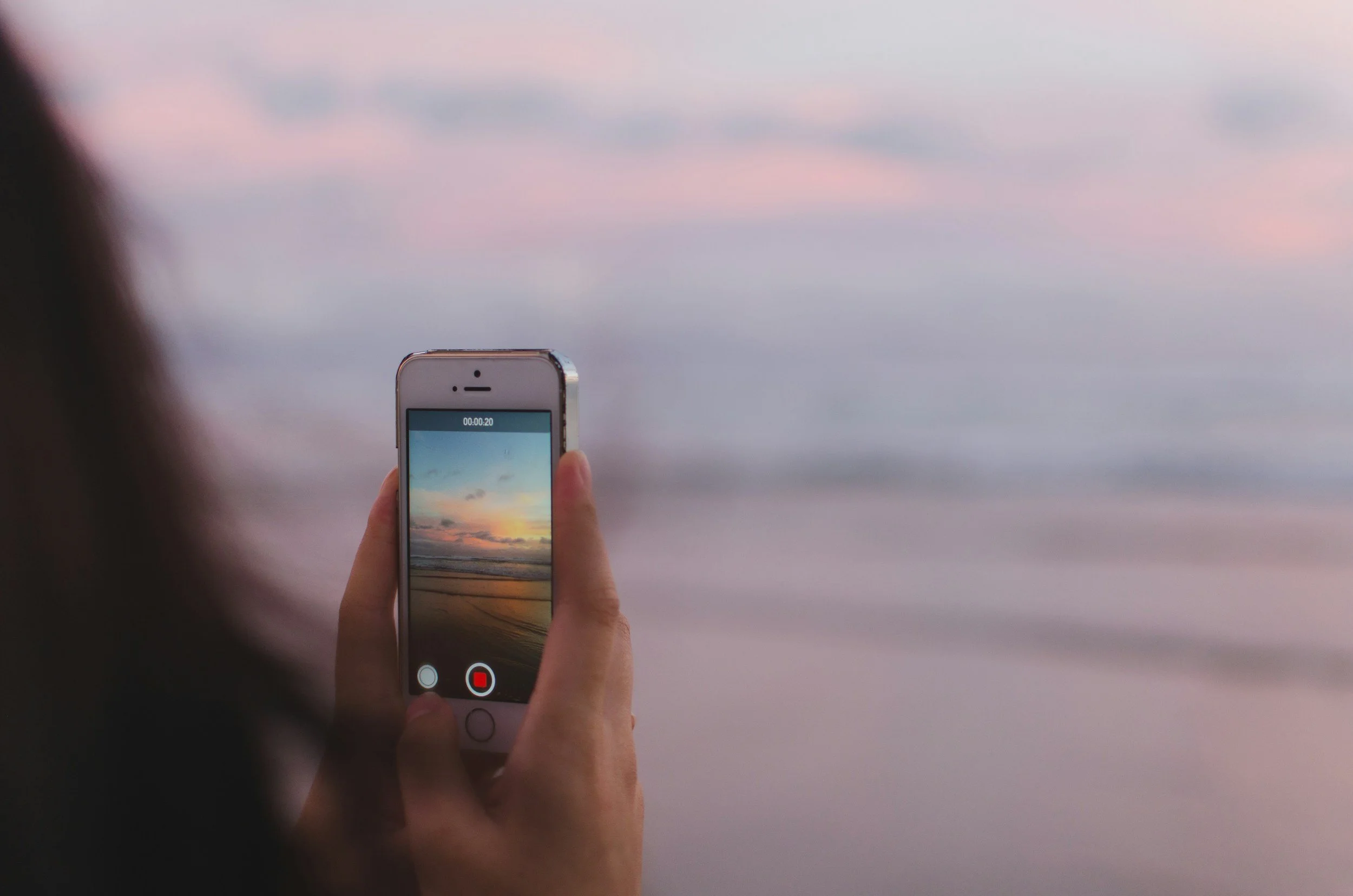 Person holding a smartphone capturing a photo or video of a sunset over the ocean with a cloudy sky and a sandy beach