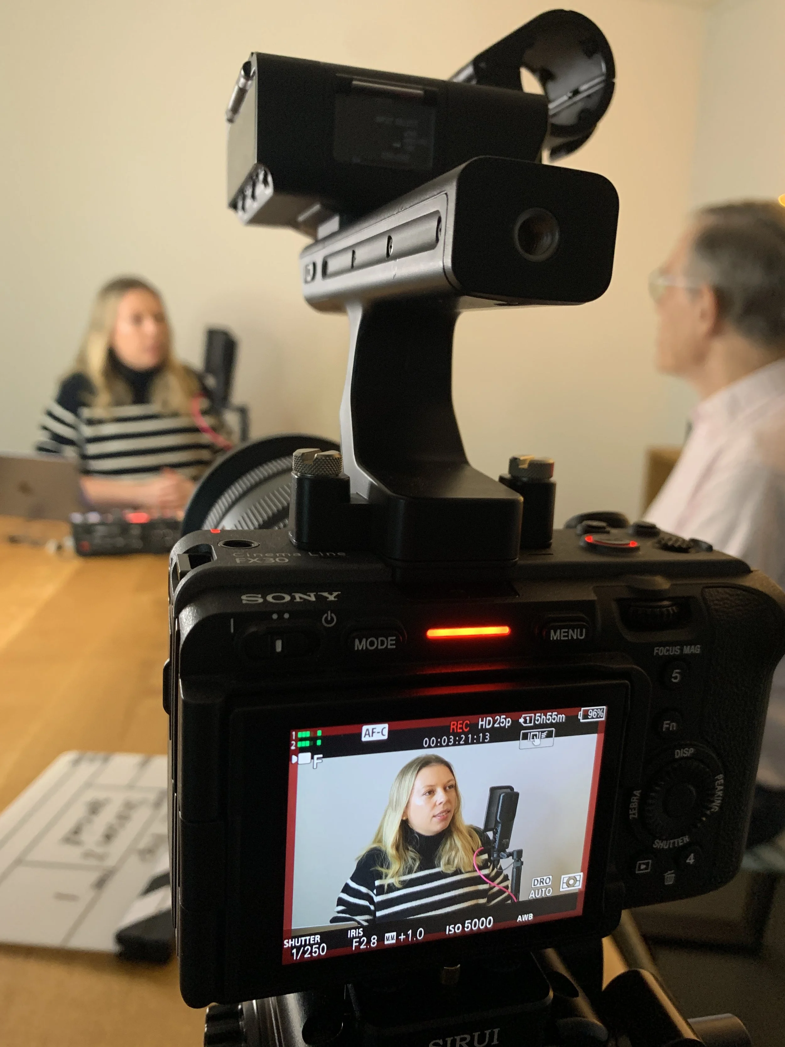 A professional camera recording a woman speaking into a microphone during an interview, with another person in the background at a table.