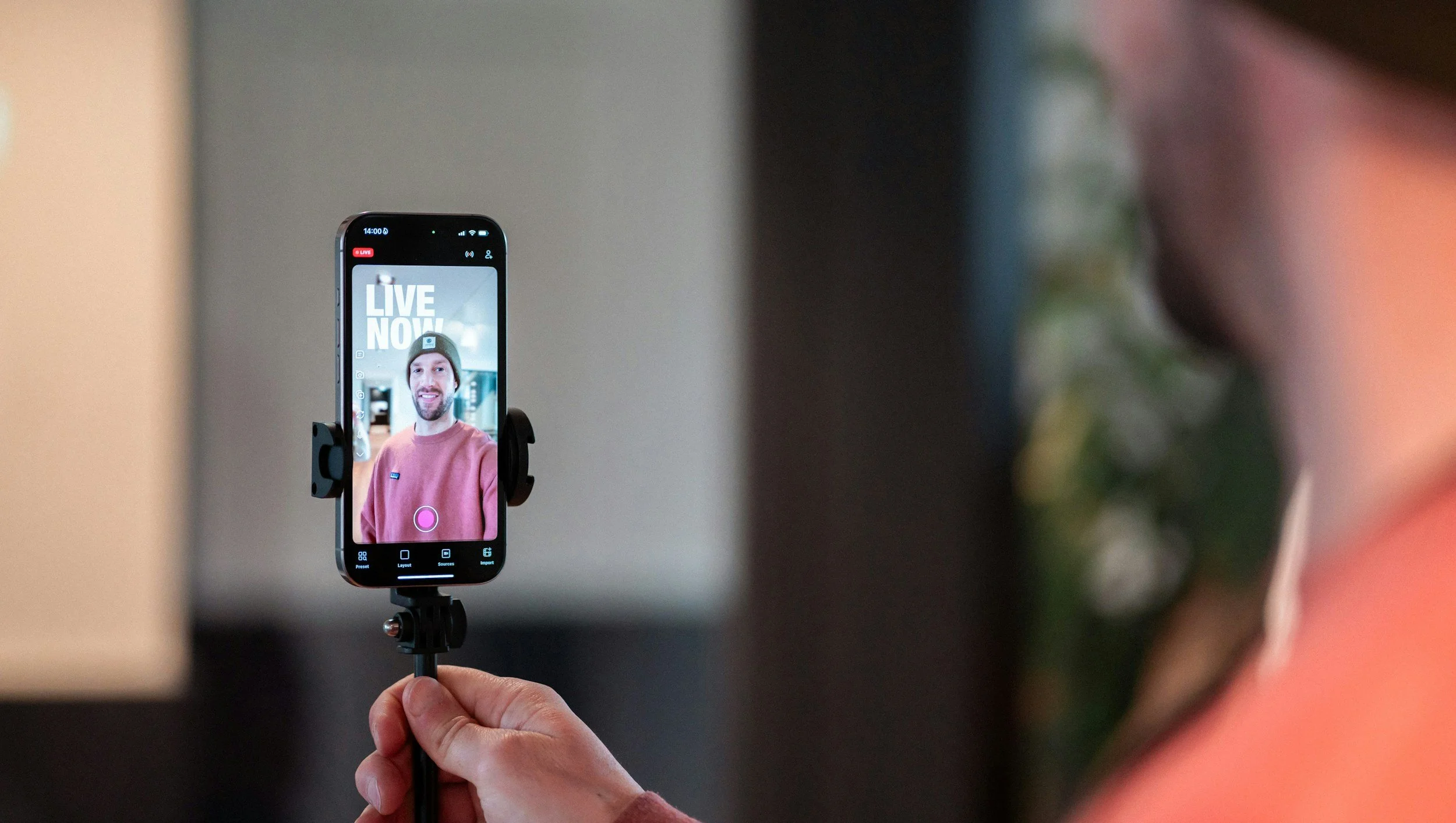 A person holding a smartphone on a tripod, recording a live video of a man wearing a pink sweatshirt and a beanie, smiling at the camera. The phone screen shows the live video with the text "LIVE NOW" in the background.