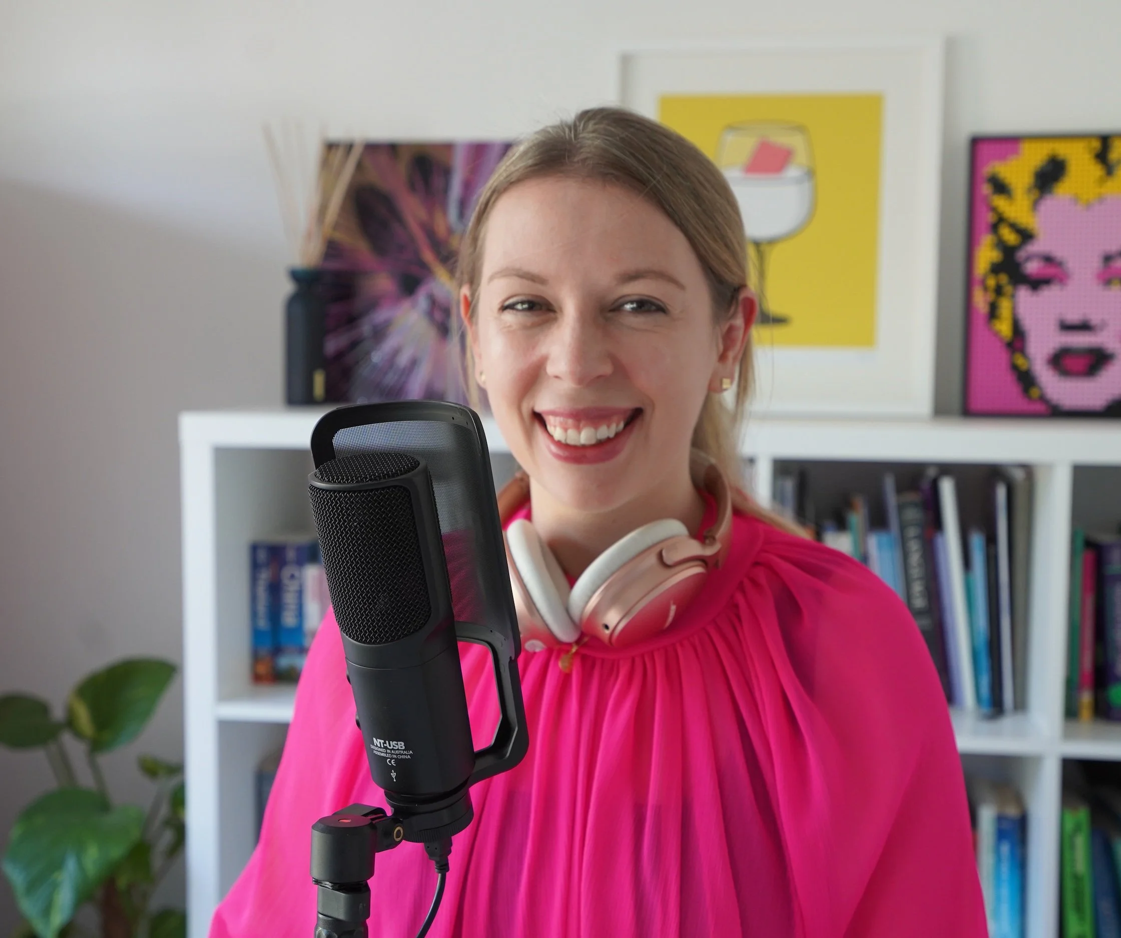 Podcast and Video Producer, Laura, is standing in front of a microphone smiling into the camera. She is wearing headphones around her neck.