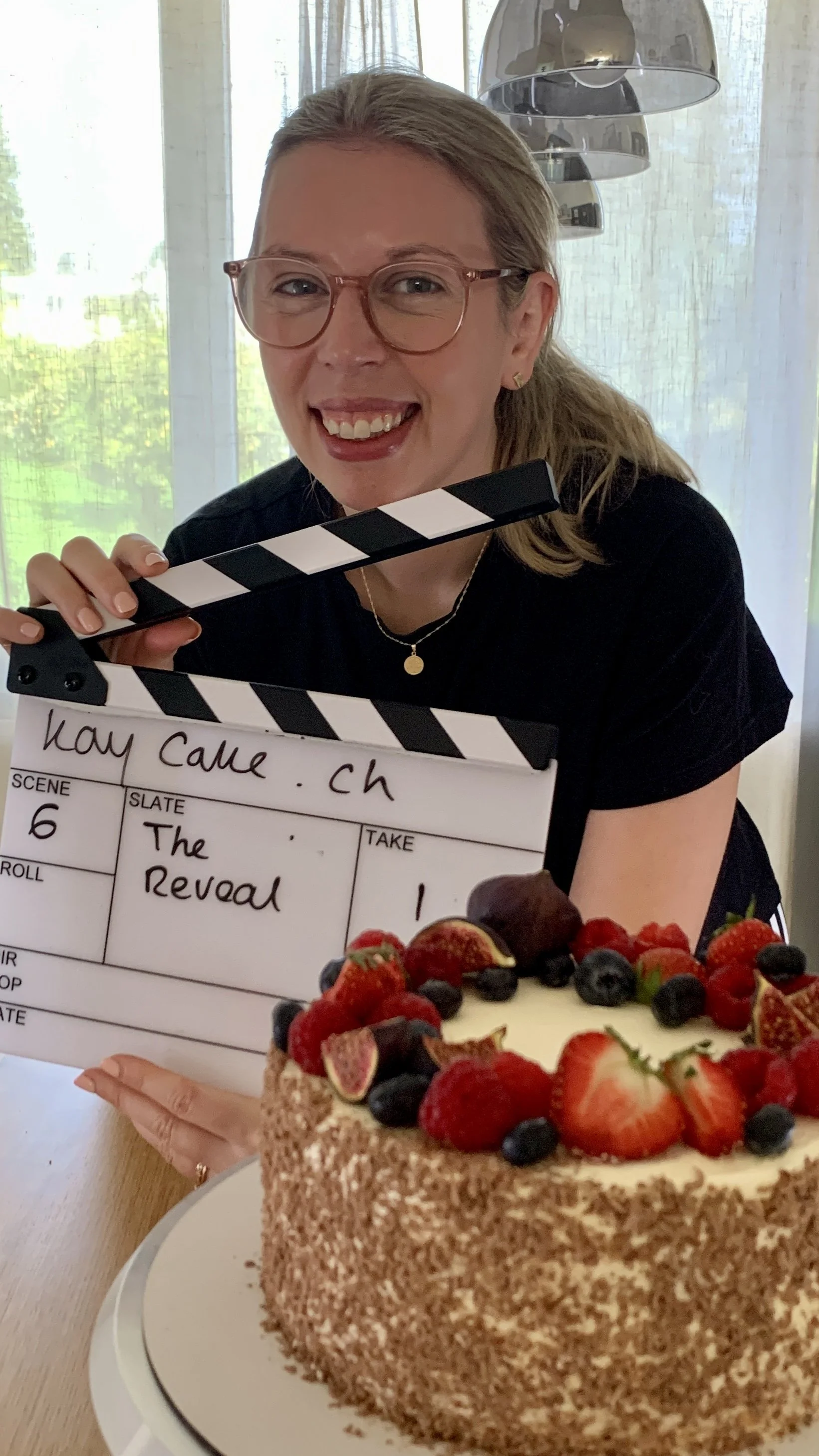 A woman with glasses holding a clapperboard and a cake with strawberries, blueberries, and other berries, celebrating a film shoot or occasion.