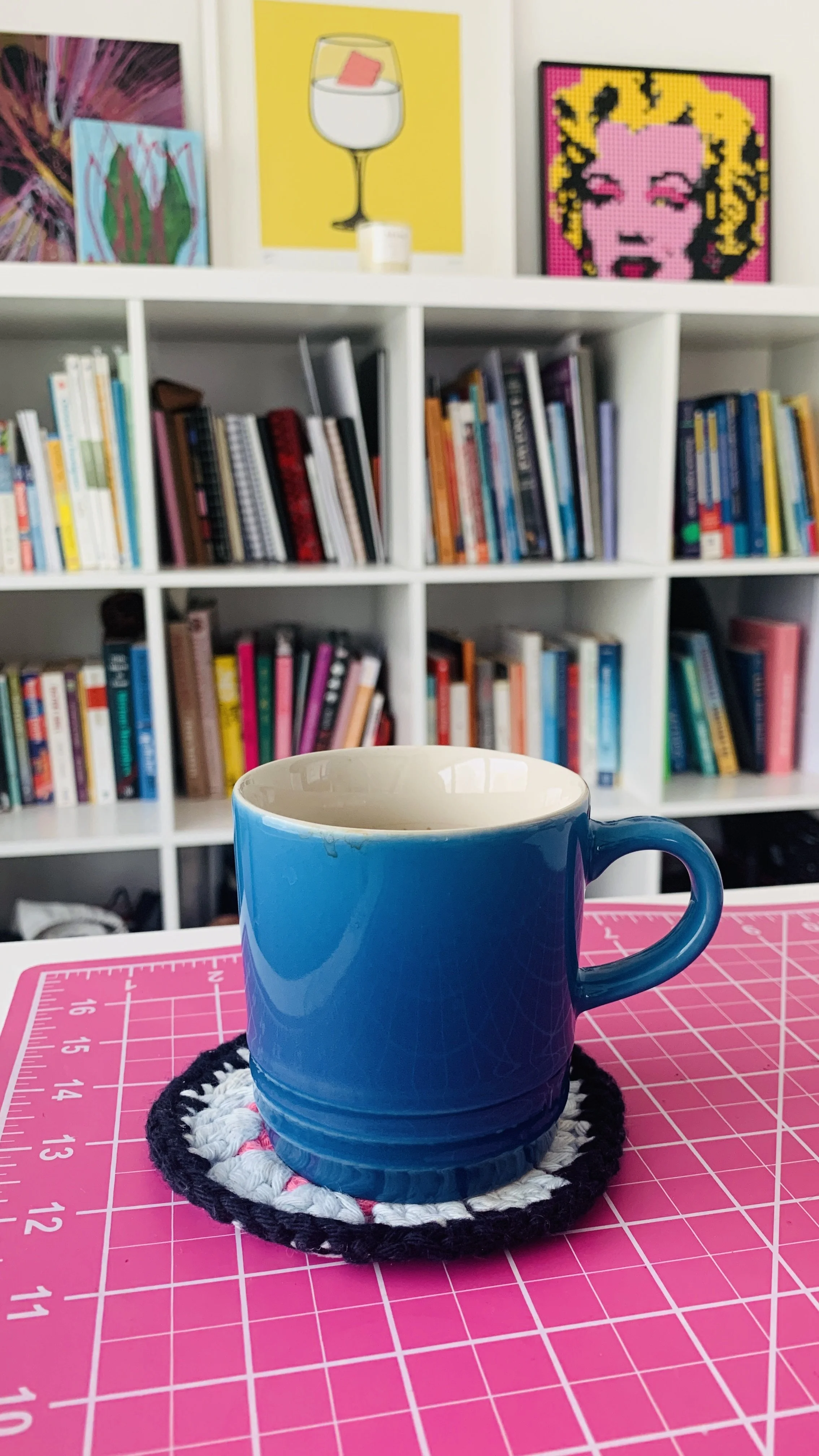 A photo of a coffee cup sat on a desk in a colourful studio. In the background, there is a bookshelf full of books and artwork.