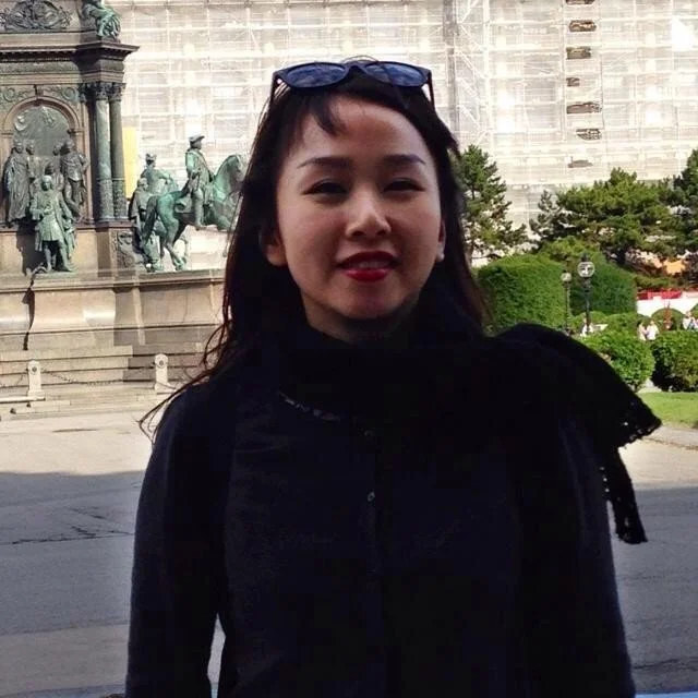Woman with dark hair and sunglasses on her head smiling outdoors in front of a historical monument and green trees.