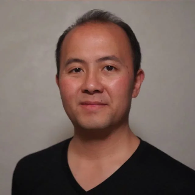 Portrait of a man with short dark hair and fair skin, wearing a black shirt, standing against a plain background.