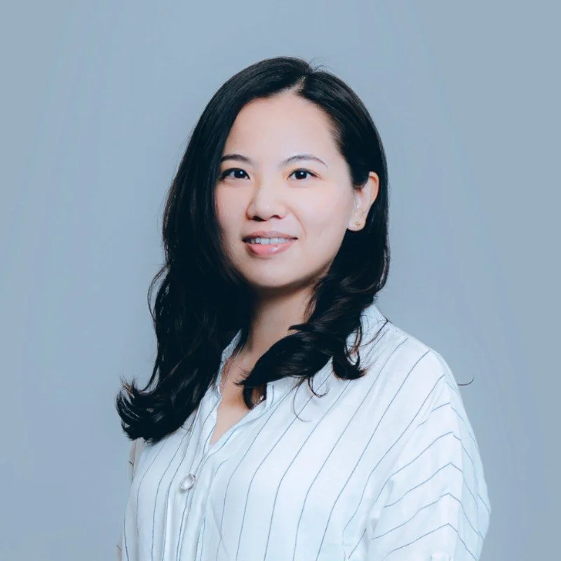 A woman with long, dark hair and light skin, wearing a white blouse with thin black stripes, smiling slightly against a plain gray background.
